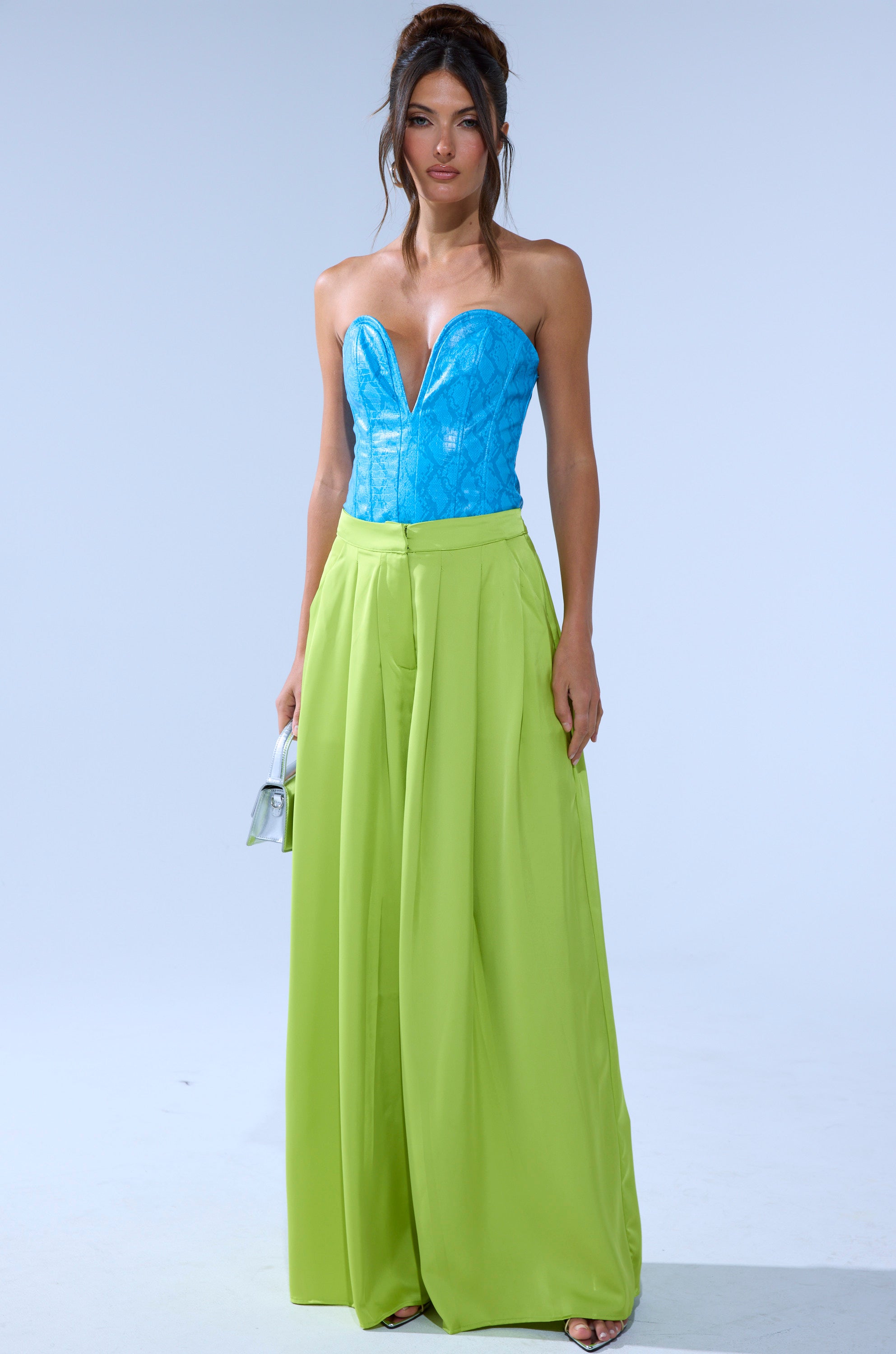 A woman poses in a turquoise strapless corset top and CHARTREUSE PLEATED PALAZZO PANT with a wide-leg, pleated design, holding a small silver handbag against a plain light background. 6