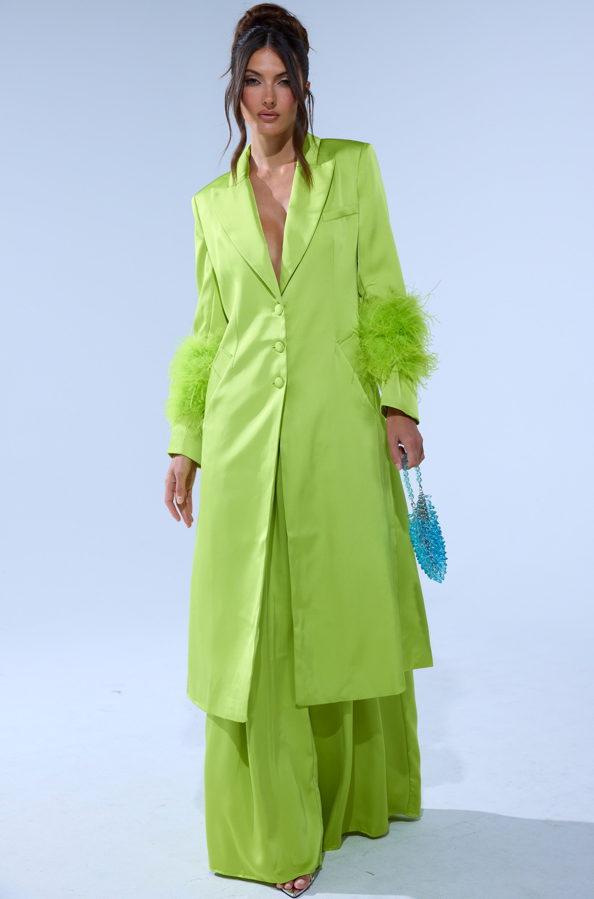 A woman wears the CHARTREUSE FEATHER TRIM TRENCH with feathery cuffs, matching wide-leg pants, open-toed shoes, and holds a small blue beaded purse against a plain light background. 0