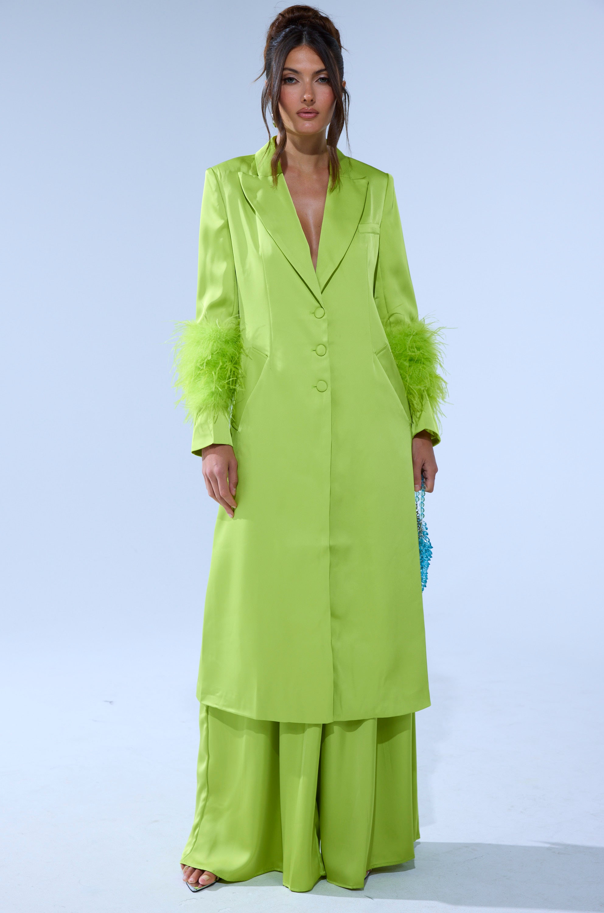 A woman models the CHARTREUSE FEATHER TRIM TRENCH in bright lime green with feathered sleeves, styled with matching wide-leg pants and open-toe shoes. Her hair is up with loose strands, set against a plain light background. 1