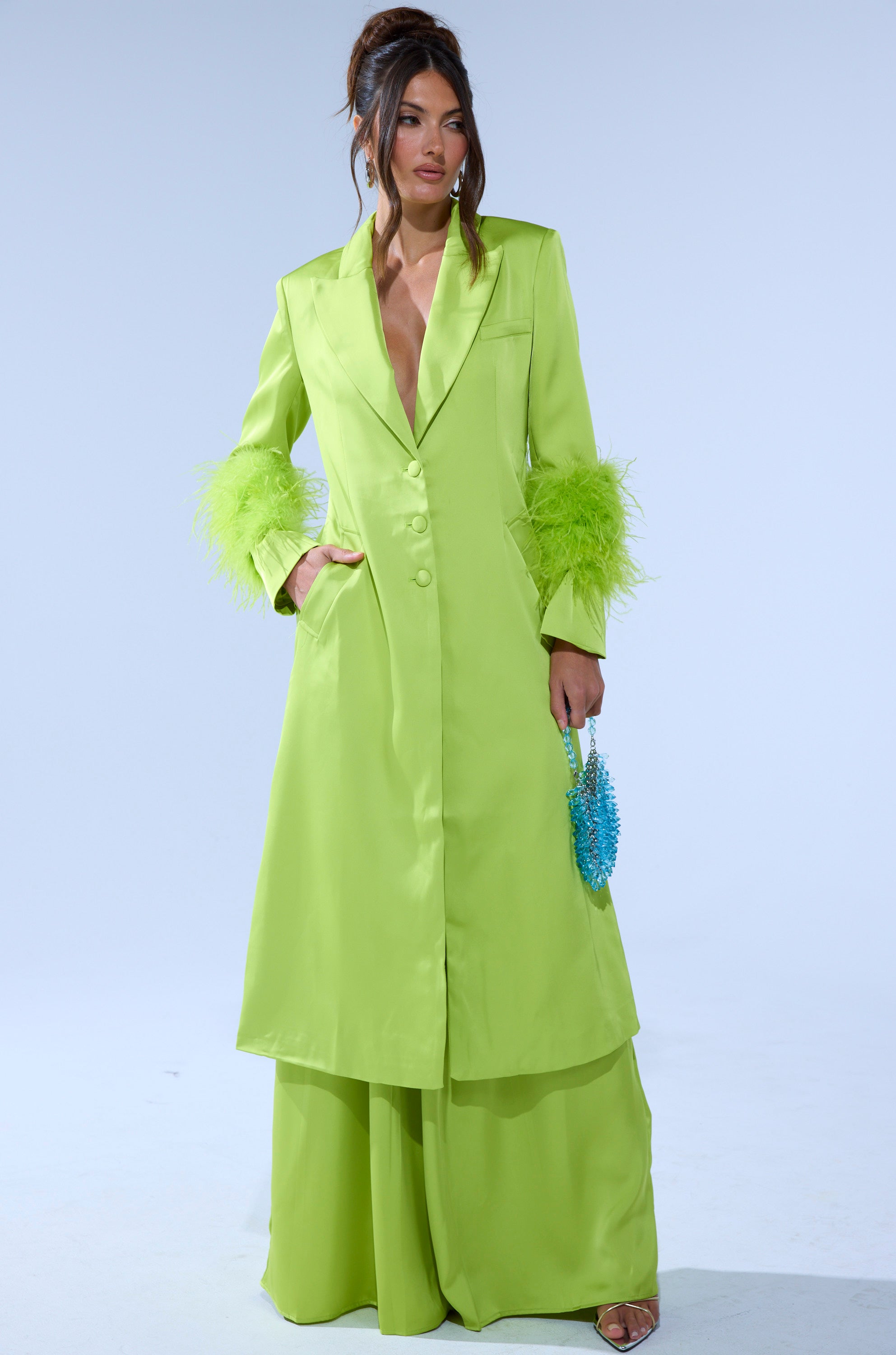 A woman poses against a plain light background wearing the CHARTREUSE FEATHER TRIM TRENCH over matching wide-leg pants and holding a small blue textured handbag. 2