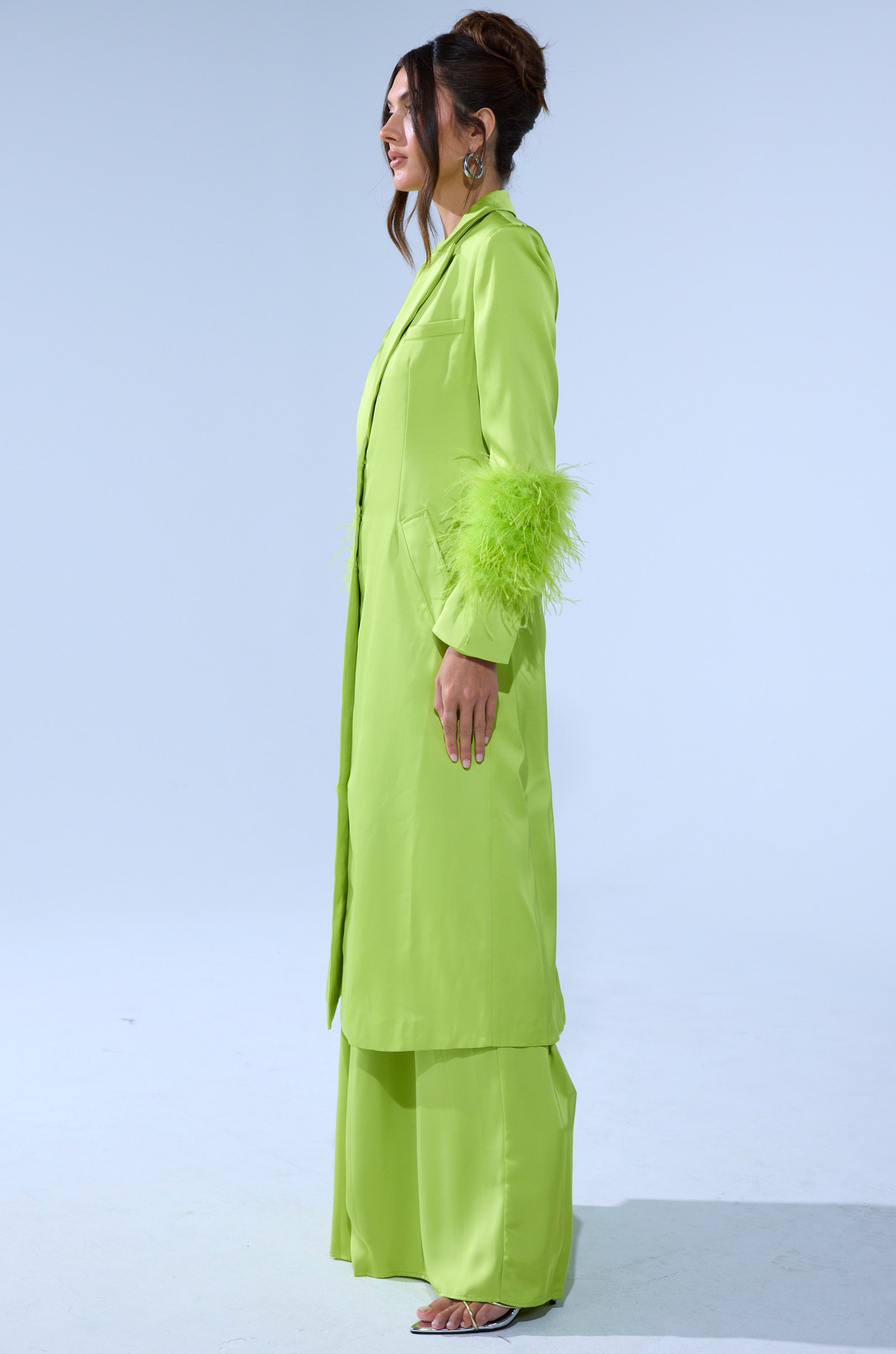 A woman stands sideways in the CHARTREUSE FEATHER TRIM TRENCH with feathered cuffs, matching wide-legged pants, and heeled sandals. Her hair is in a loose bun and she wears hoop earrings against a plain light background. 3