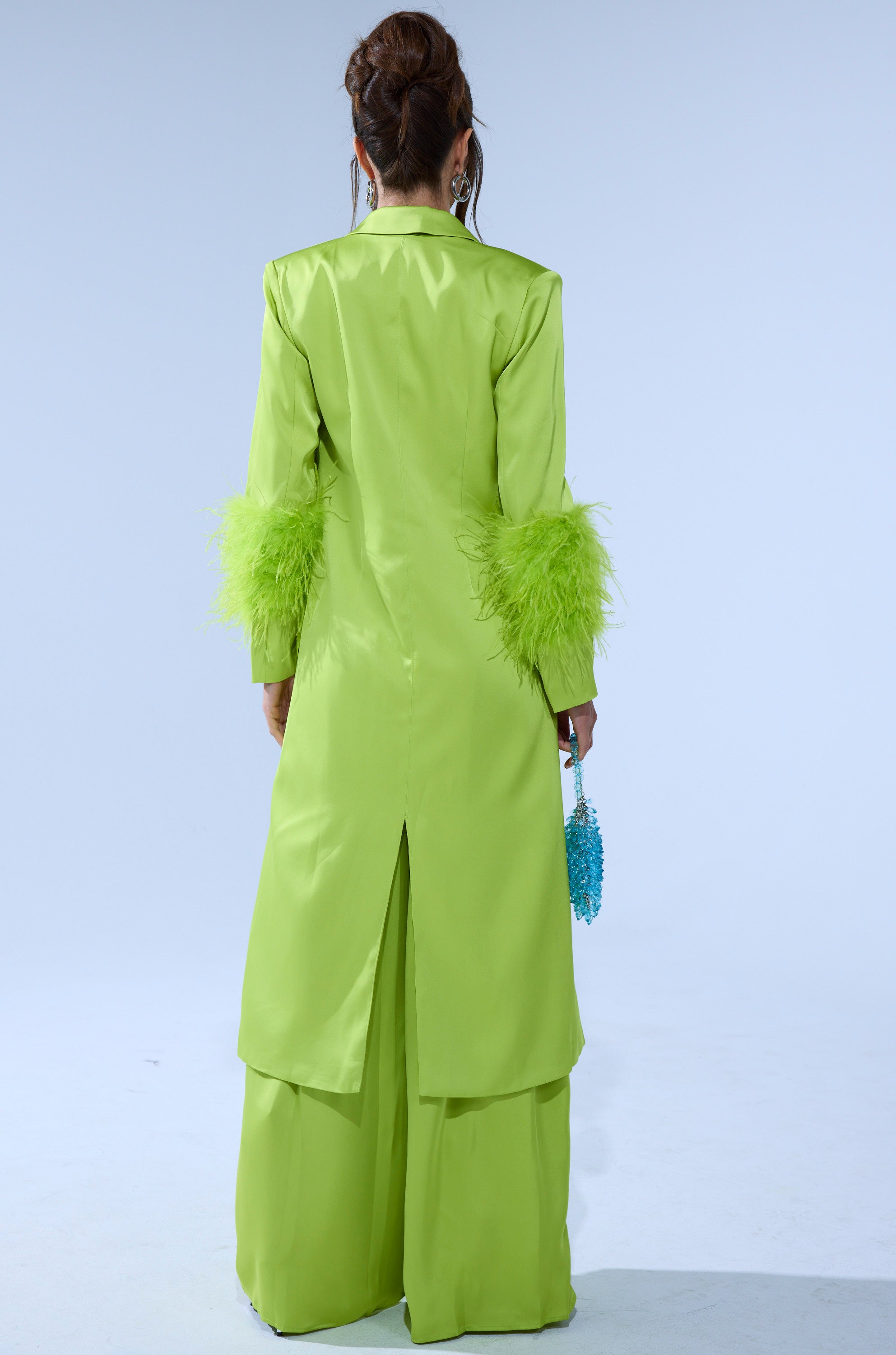 A person with long brown hair tied up stands facing away, wearing the CHARTREUSE FEATHER TRIM TRENCH and wide-legged pants, holding a small blue beaded purse against a plain light background. 4