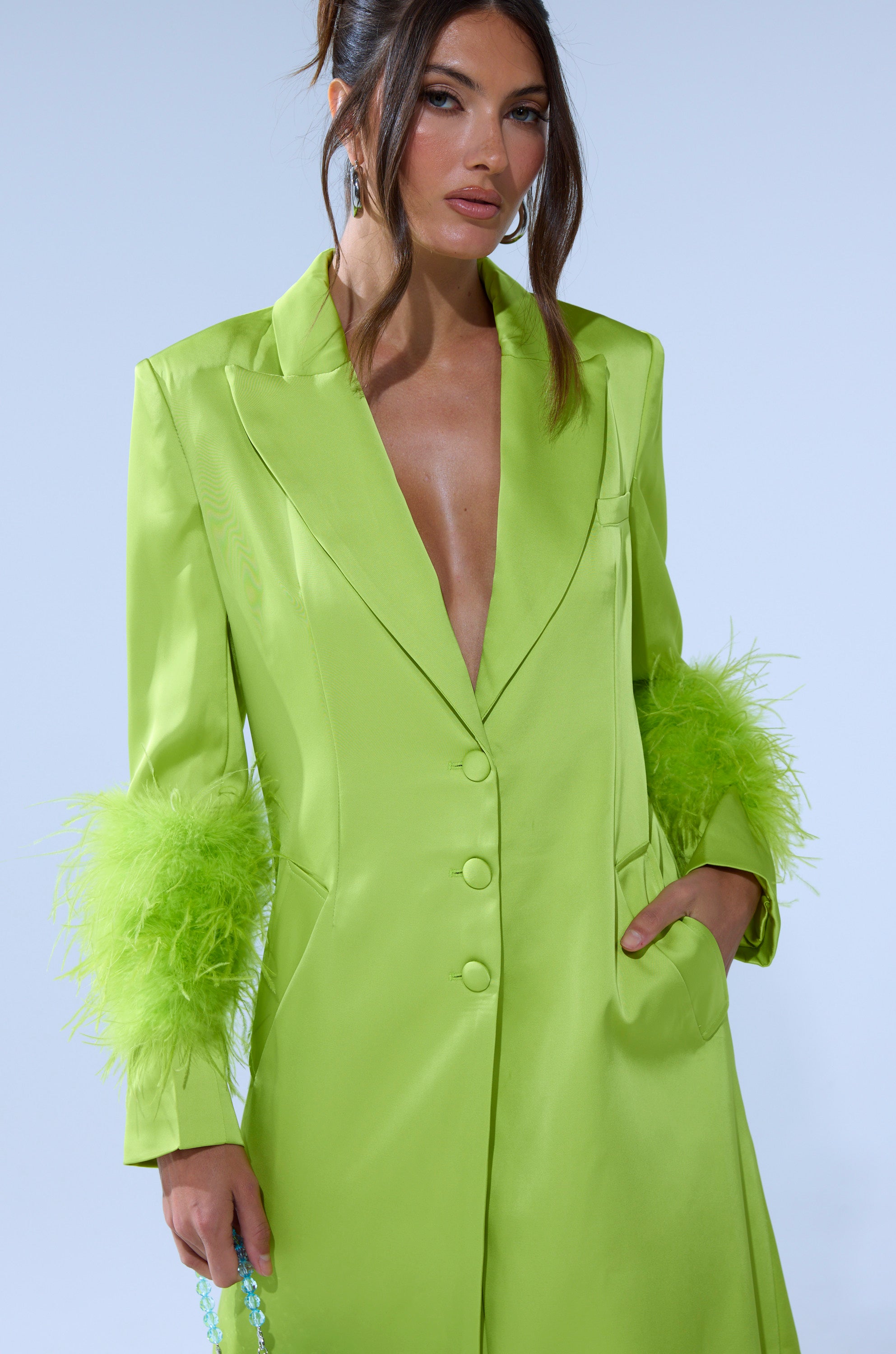 A woman models the CHARTREUSE FEATHER TRIM TRENCH, a neon green longline jacket with feathered cuffs, posing with one hand in her pocket against a plain light background. 5
