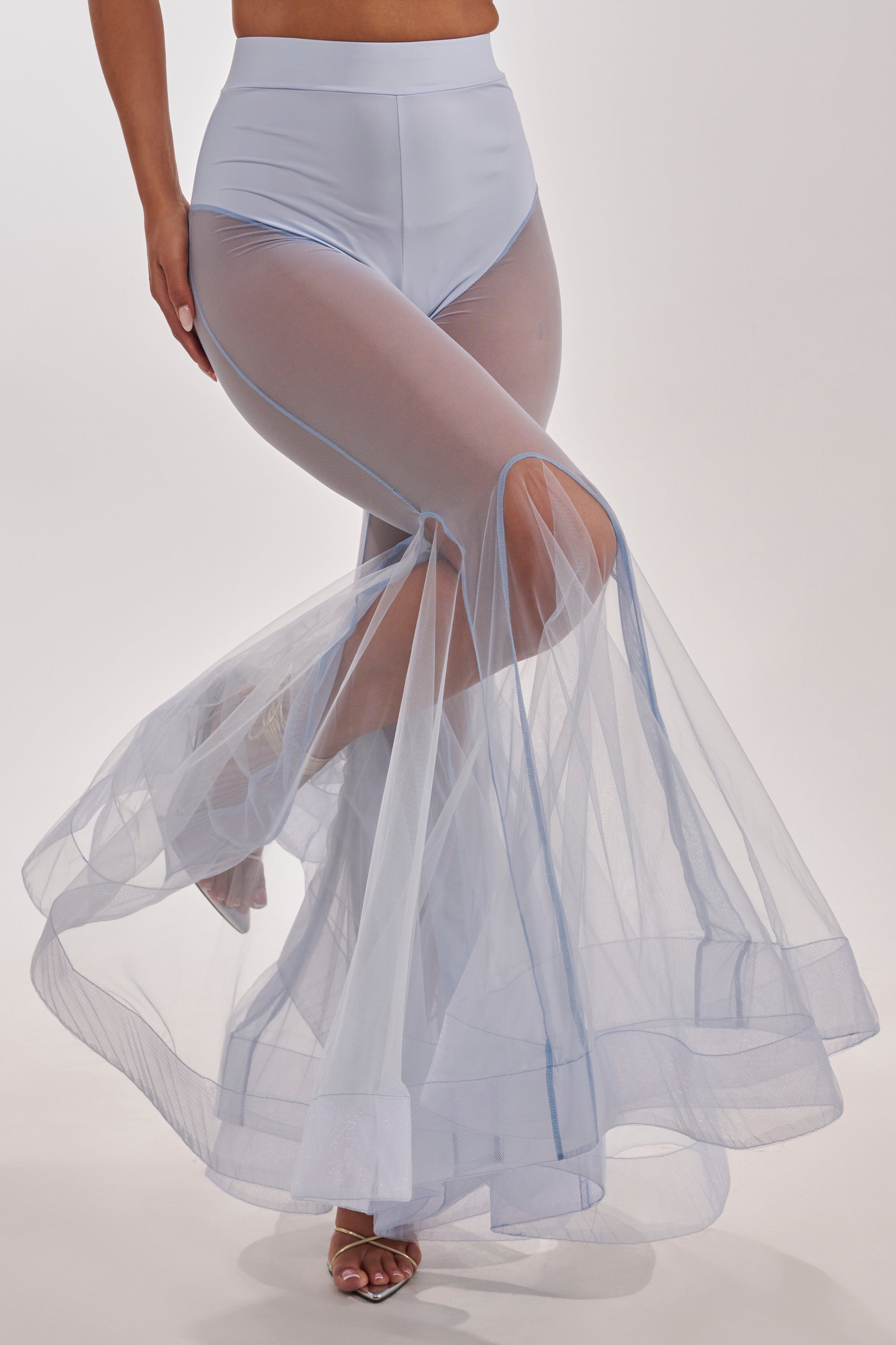 A person wearing the FEELING BLUE MESH FLARE PANT, featuring sheer, light blue fabric with a mermaid-inspired flare silhouette, poses with one leg forward and open-toe heels. 3