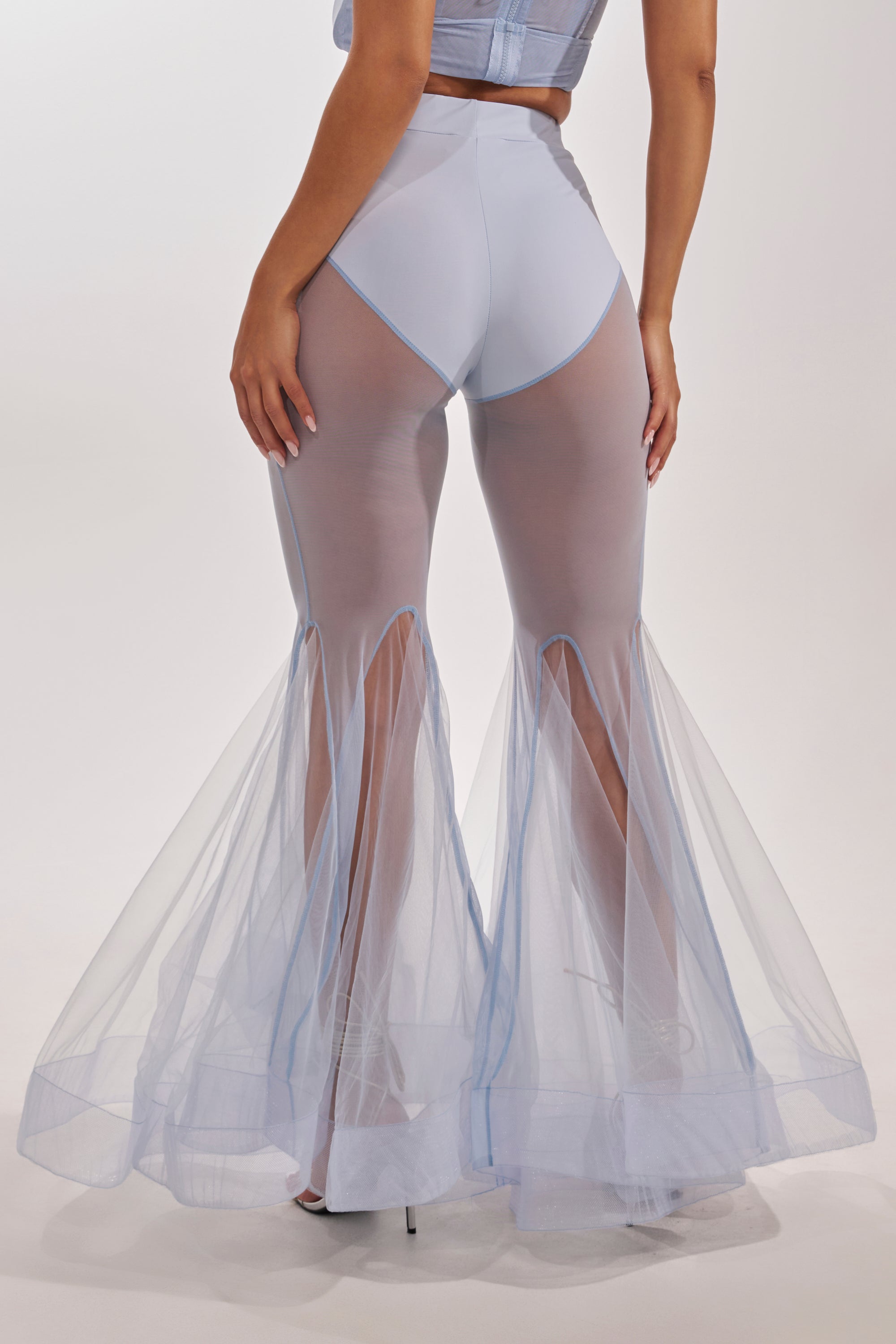 A woman stands with her back to the camera in FEELING BLUE MESH FLARE PANT, featuring high-waisted blue mesh and flared tulle-like panels from the knee down, creating a mermaid-inspired silhouette against a plain background. 5