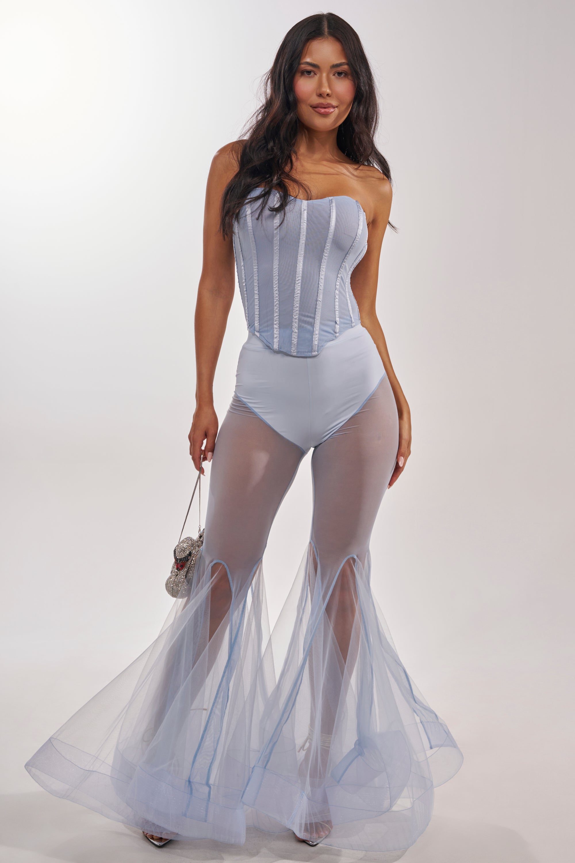 A woman models the FEELING BLUE MESH FLARE PANT—high-waisted, blue mesh pants with a mermaid-inspired flare silhouette—paired with a light blue corset bodysuit. She stands against a white background, smiling with her hair down and holding a small purse. 6