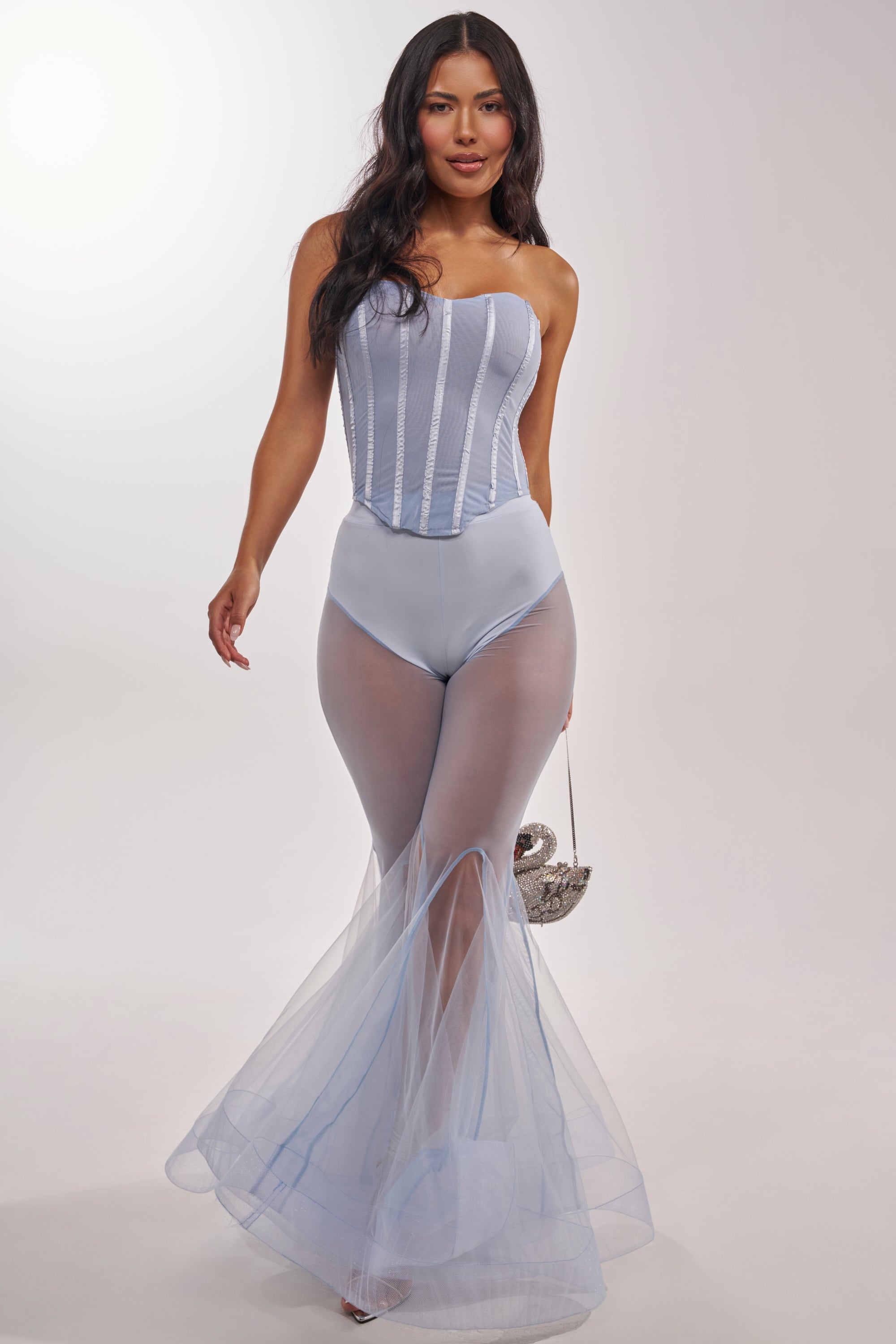 A woman wearing a strapless, light blue corset bodysuit and the FEELING BLUE MESH FLARE PANT, paired with a small silver swan-shaped handbag, stands against a plain light background. 7