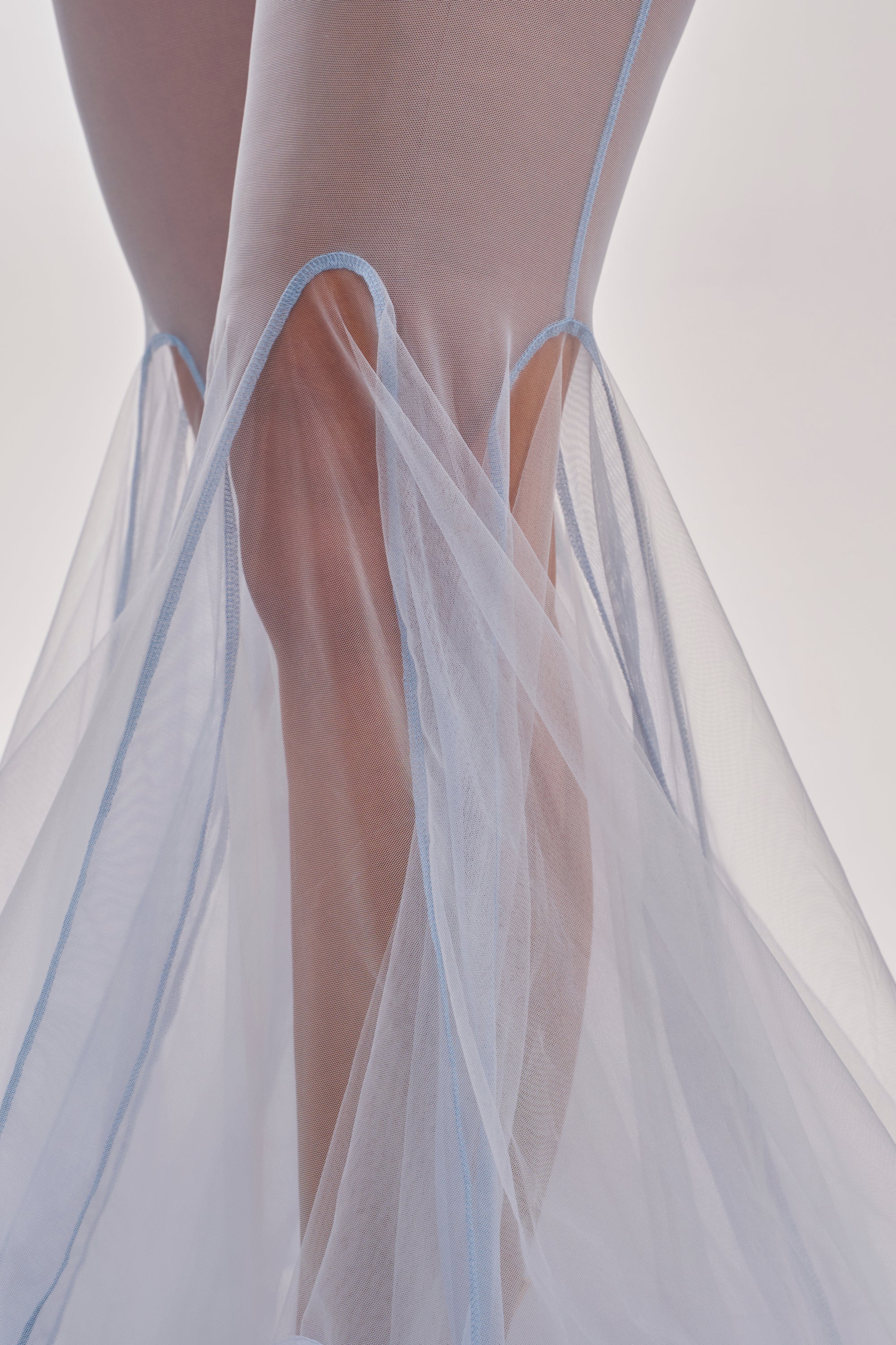 Close-up of a person’s legs in the FEELING BLUE MESH FLARE PANT, featuring blue sheer mesh with light blue seams and a mermaid-inspired silhouette that creates a soft, airy effect against a light background. 9