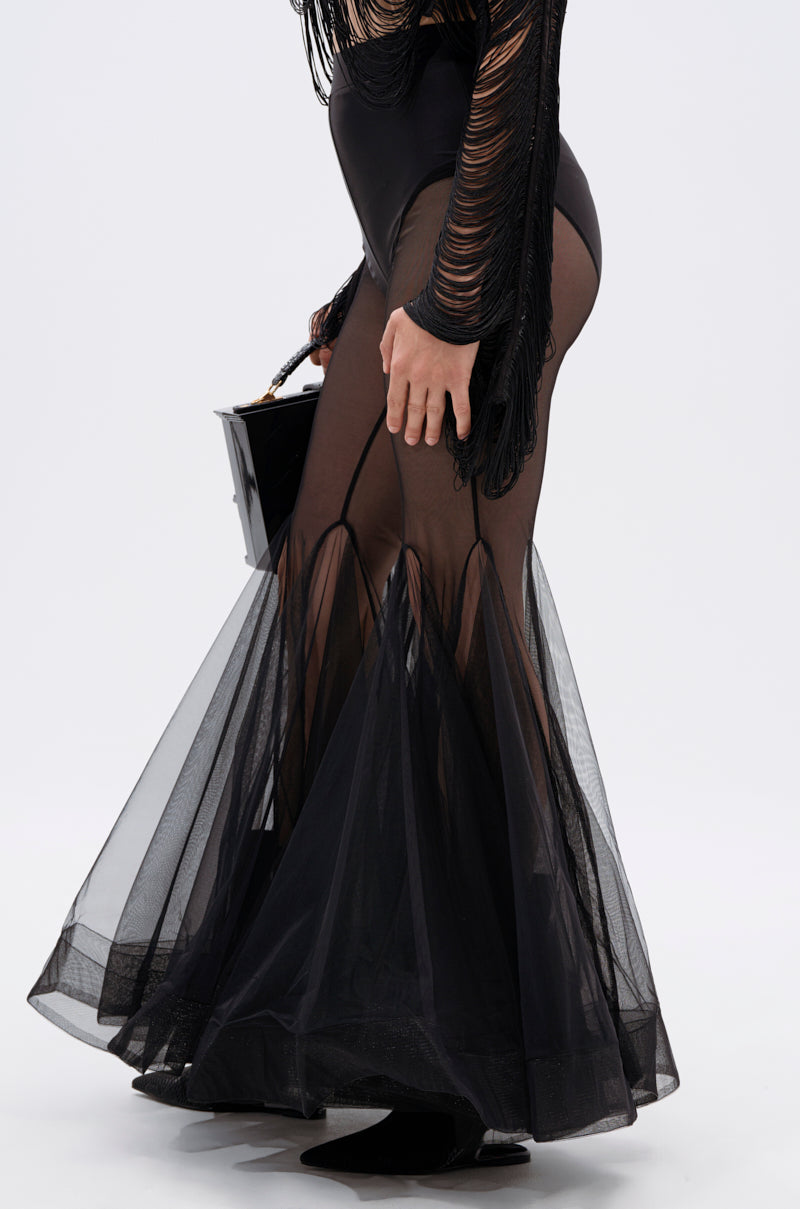 A person wears the IT ONLY GETS BETTER MESH FLARE LEGGING—sheer black mesh leggings with layered flares and solid panels—paired with a shiny black handbag, standing against a plain white background. 4