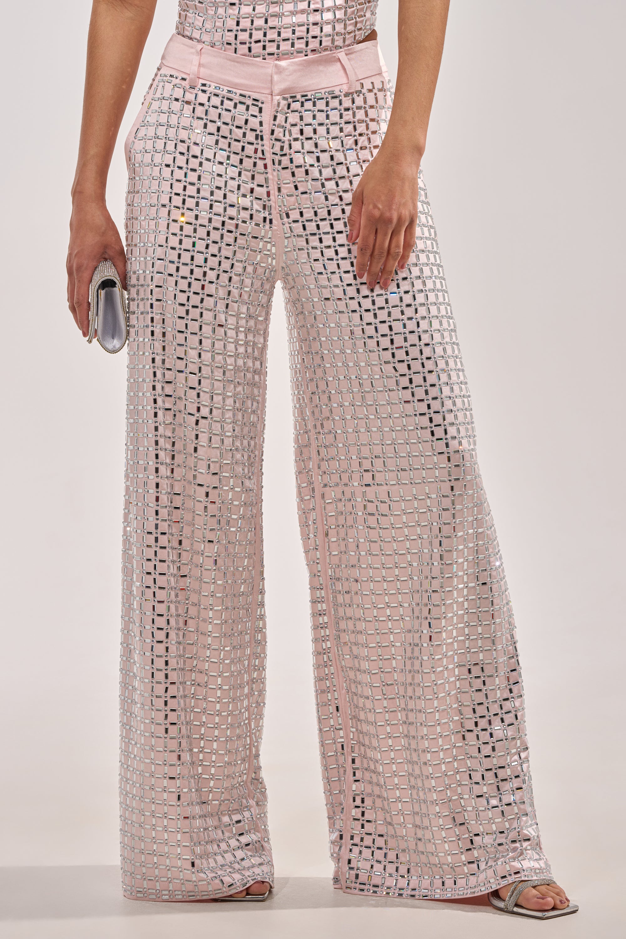 A woman wears the LILA EMBELLISHED SATIN TROUSER IN LIGHT PINK, featuring a wide-leg cut, semi-sheer satin, and rhinestone embellishments in a silver grid. She pairs them with heeled sandals and a small silver clutch for a striking look. 0