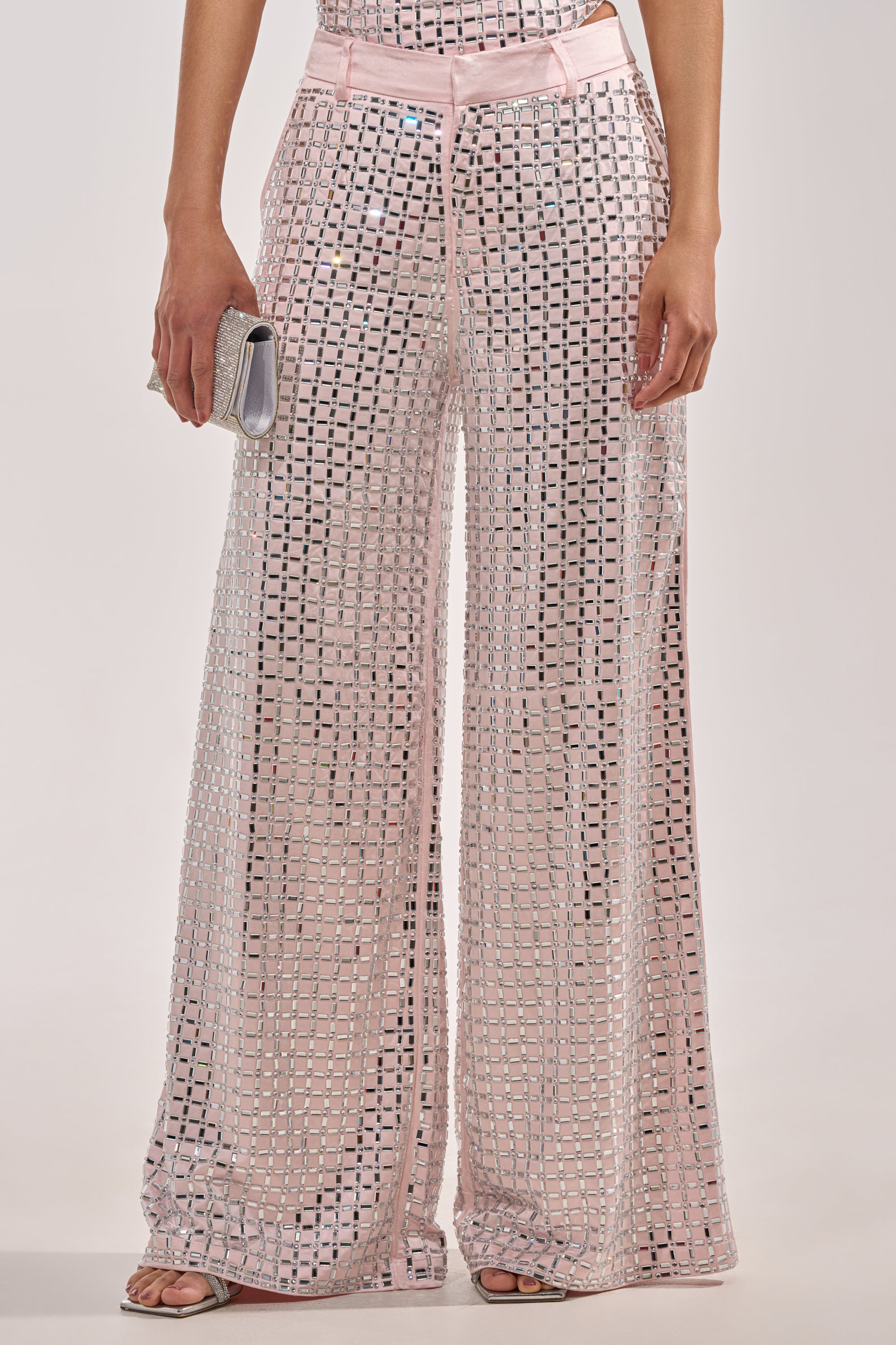 A woman wears the LILA EMBELLISHED SATIN TROUSER IN LIGHT PINK—wide-leg satin pants with sequin details—styled with open-toe heels and a small silver clutch. 2