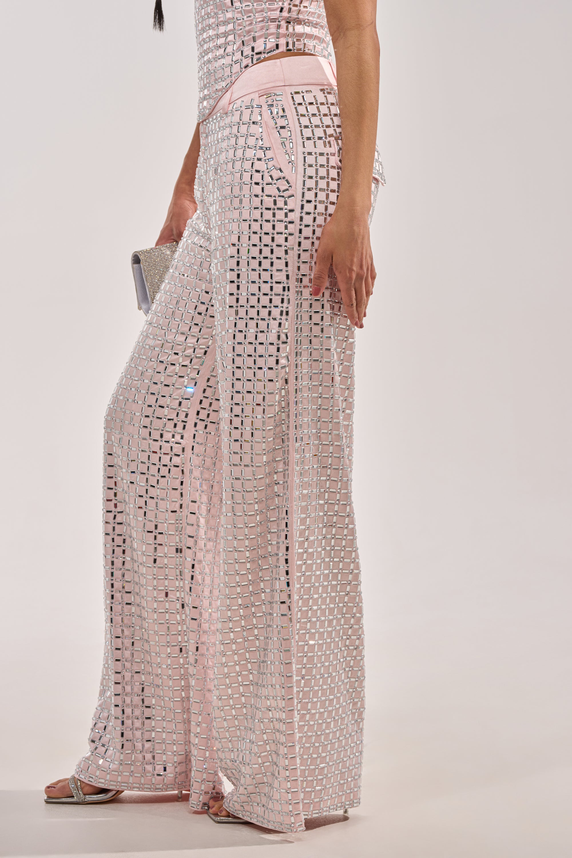 A woman wears the LILA EMBELLISHED SATIN TROUSER in light pink—high-waisted with square sequins—paired with a sparkling clutch and silver sandals against a plain light gray background. 3
