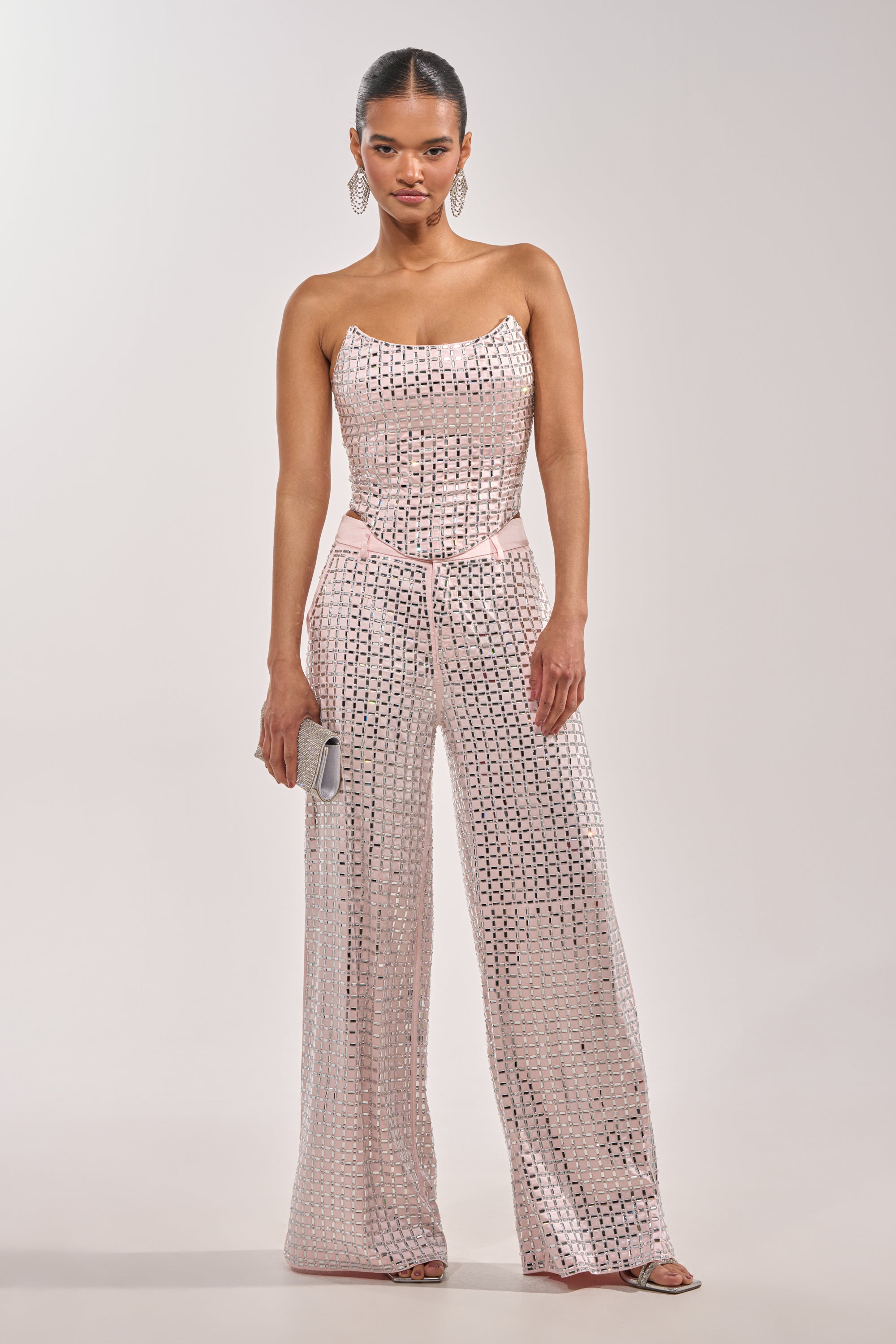 A woman stands in light pink LILA EMBELLISHED SATIN TROUSER with wide legs, a straight neckline top, silver clutch, large hoop earrings, her hair pulled back, and silver heels. 8
