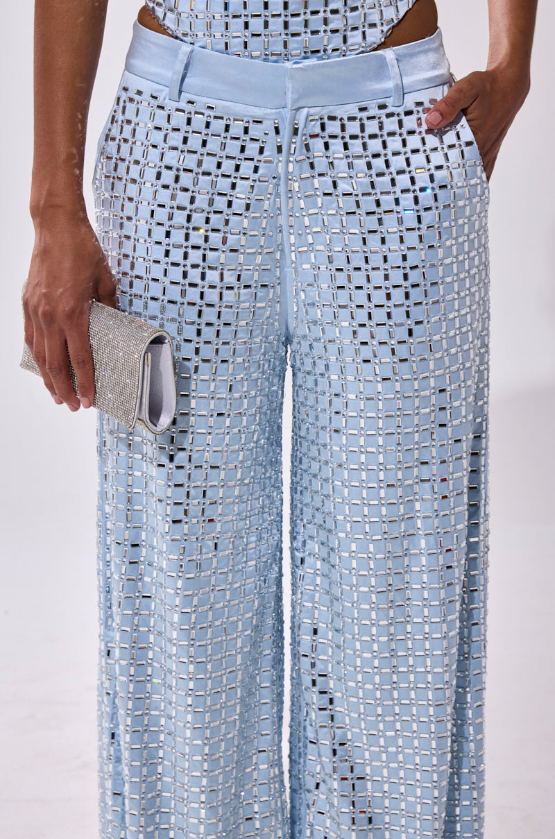 A person wears the LILA EMBELLISHED SATIN TROUSER in powder blue, featuring wide legs and subtle rhinestone details, paired with a matching top and holding a sparkly silver clutch for added glamour.