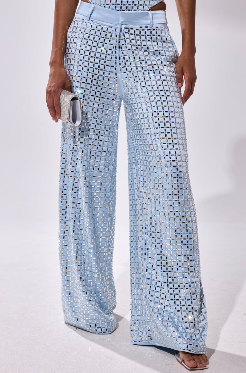 Someone wearing the LILA EMBELLISHED SATIN TROUSER in powder blue—high-waisted, wide-leg, featuring a square cutout geometric pattern over a matching underlayer—paired with a silver clutch and white open-toe heels.