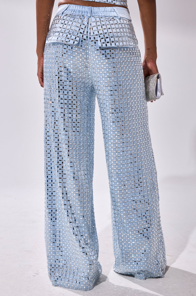 A person stands facing away, wearing powder blue LILA EMBELLISHED SATIN TROUSERS with wide legs, large back pockets, a matching belt, mesh fabric featuring square cutouts, and holding a small silver clutch.