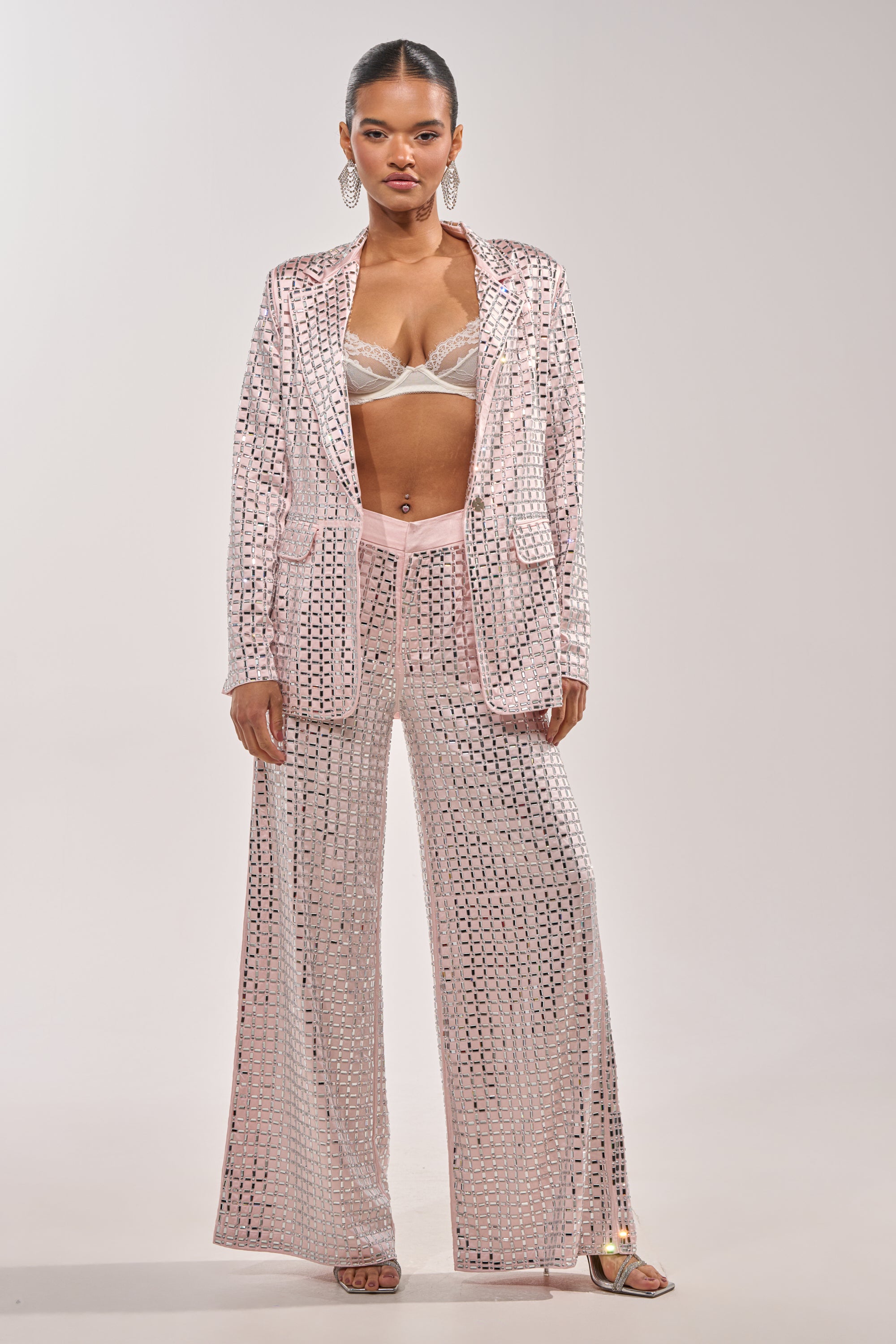 A woman wears the LILA EMBELLISHED SATIN BLAZER IN LIGHT PINK with wide-leg pants, a jeweled bralette, slicked-back hair, statement earrings, and silver open-toe heels. 9