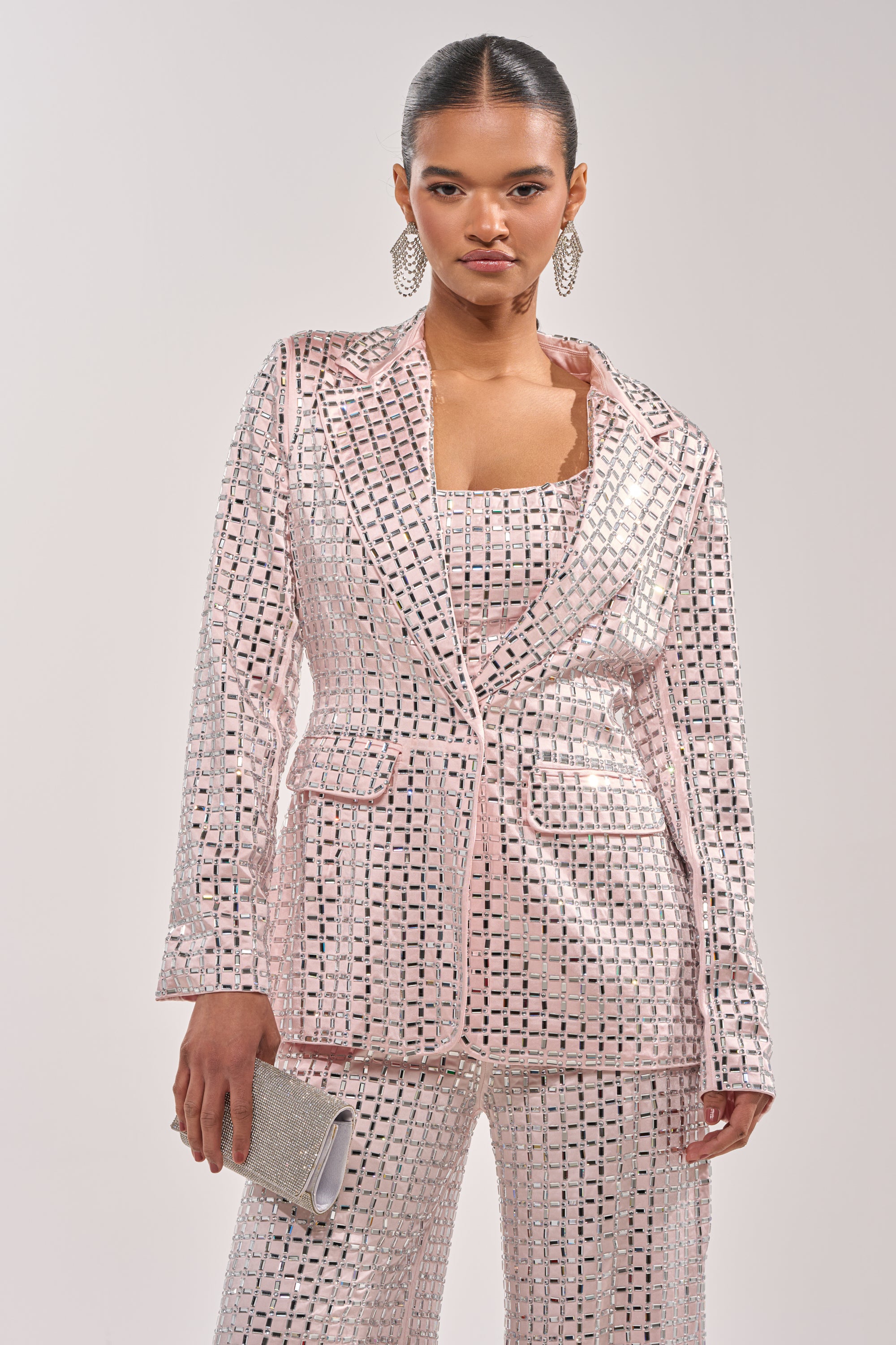 A woman wears the LILA Embellished Satin Blazer in Light Pink as a pantsuit with a matching top, paired with a silver clutch. She sports slicked-back hair, large dangling earrings, and stands against a plain light background. 1