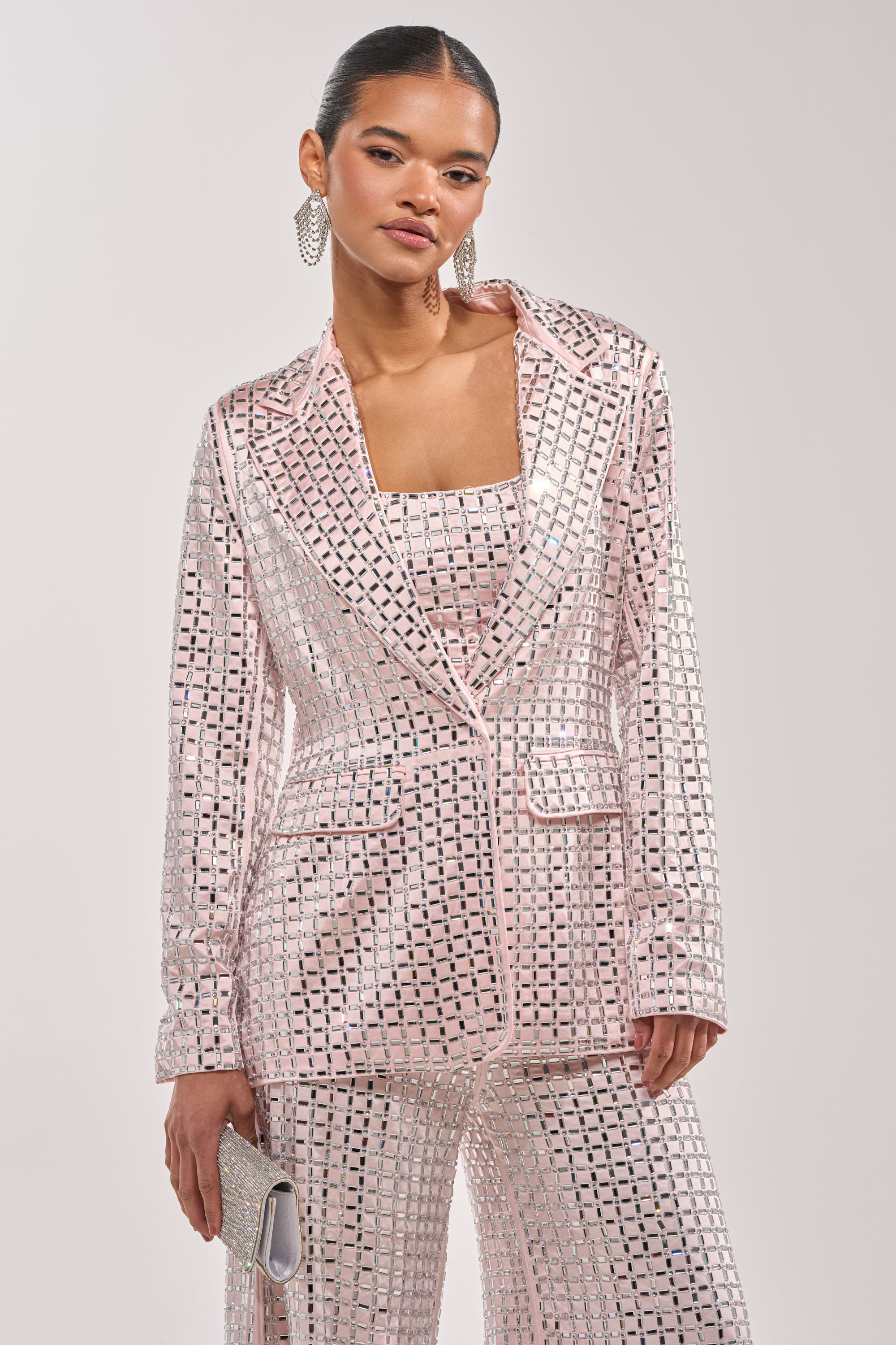 A woman models the LILA EMBELLISHED SATIN BLAZER IN LIGHT PINK, a tailored, rhinestone-studded party blazer. She pairs it with a silver clutch and statement earrings, her hair slicked back against a plain background. 2