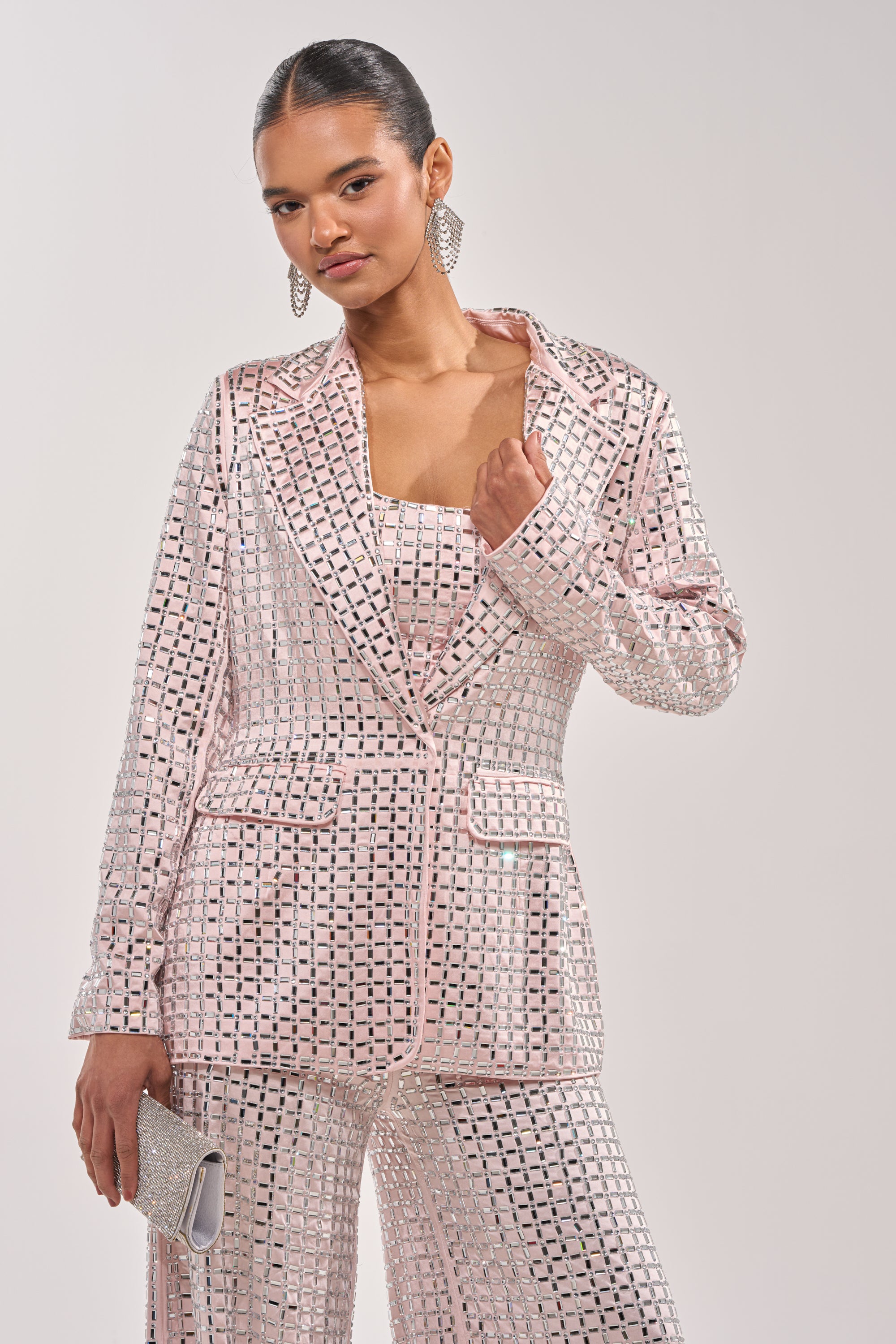 A woman stands confidently in the LILA EMBELLISHED SATIN BLAZER IN LIGHT PINK, paired with sequined pants and a silver clutch. She wears her hair slicked back and accessorizes with large geometric earrings. 3