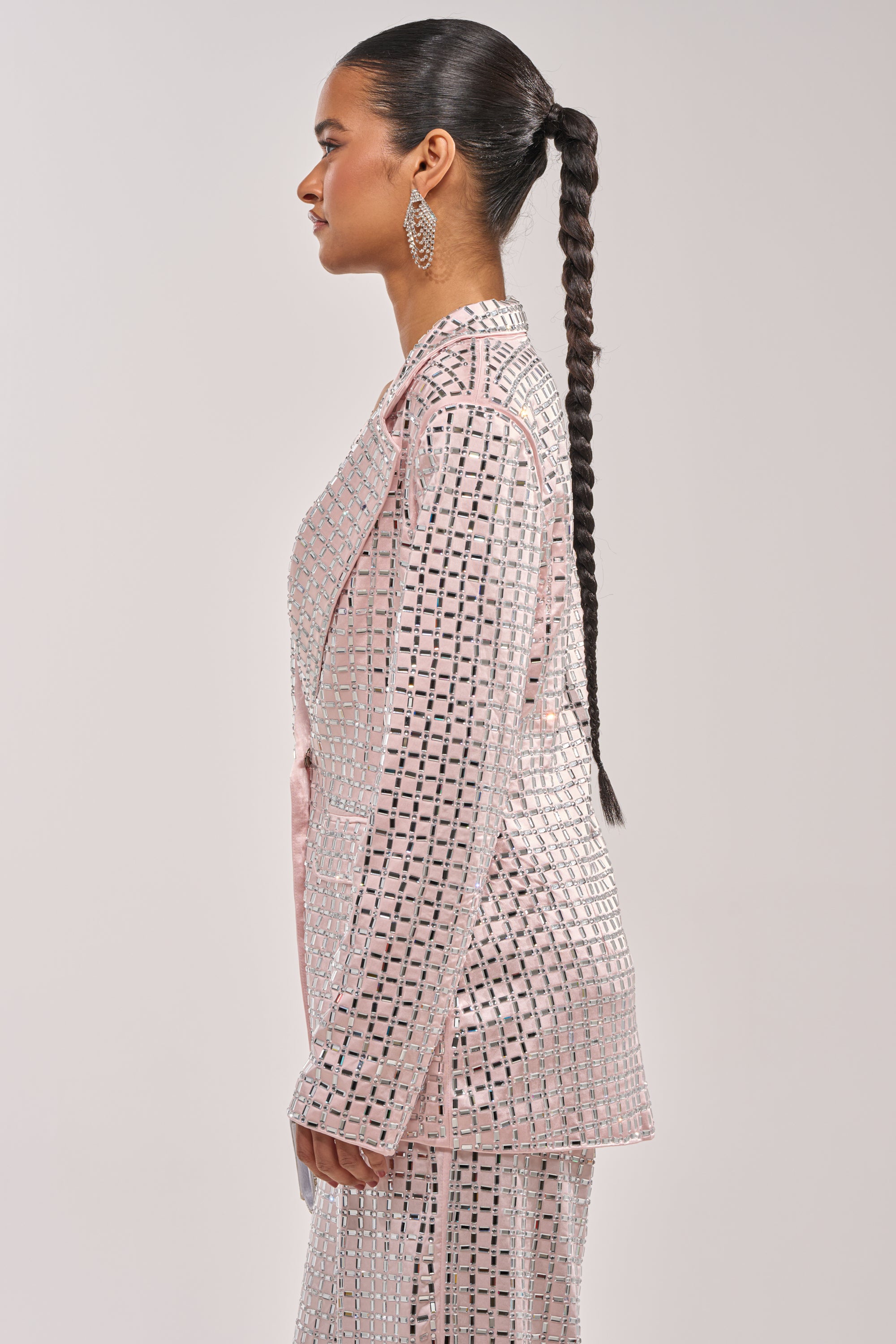 A woman with a long braided ponytail wears the LILA EMBELLISHED SATIN BLAZER IN LIGHT PINK and large statement earrings, standing in profile against a plain light background. 5
