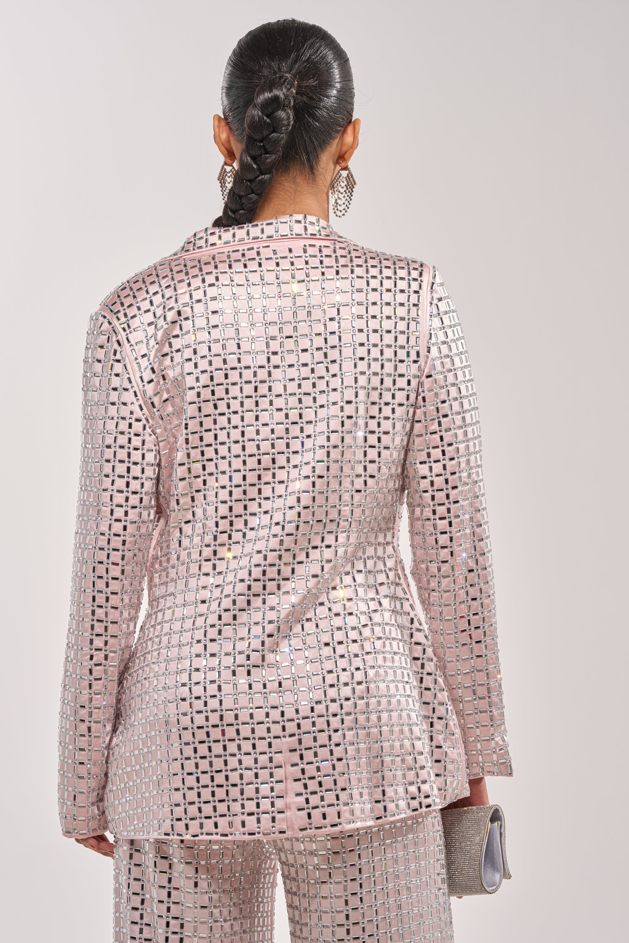 A person with dark braided hair stands facing away, wearing the LILA EMBELLISHED SATIN BLAZER IN LIGHT PINK with matching pants, accessorized with a small clutch and large sparkly earrings. 6