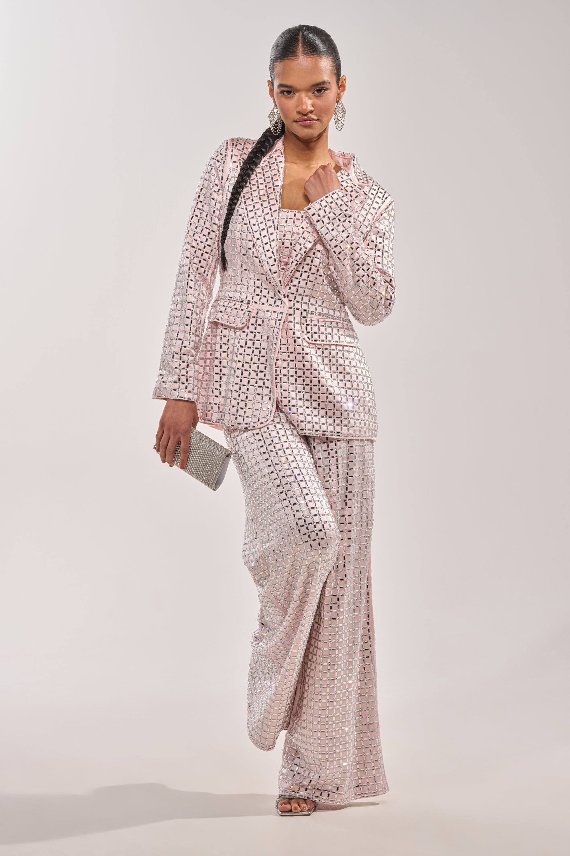 A woman stands confidently in the LILA Embellished Satin Blazer in Light Pink with matching wide-leg trousers, holding a silver clutch. Sleek braided hair and open-toed heels complete her chic party look against a plain backdrop. 8