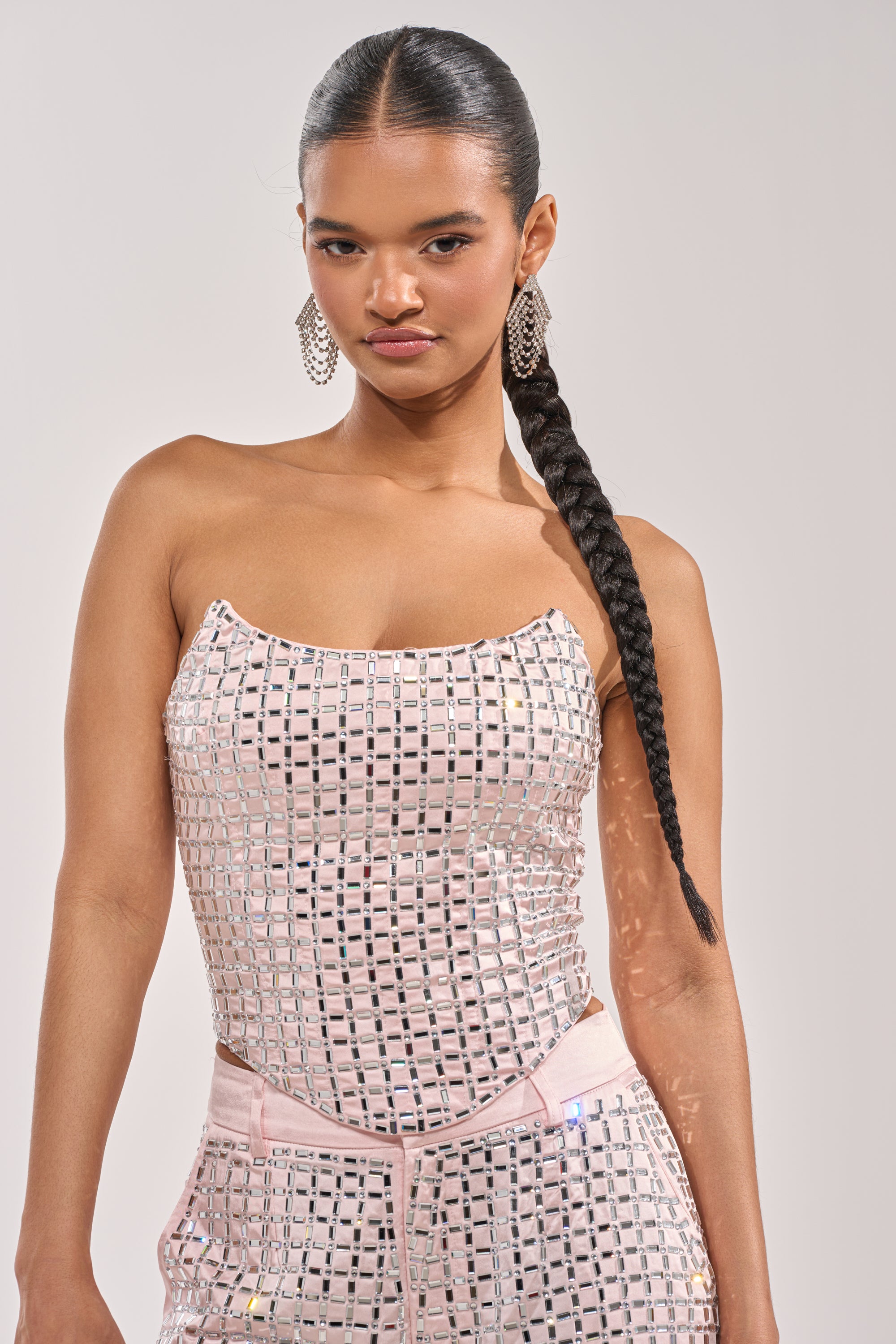 A woman with a long braided ponytail wears the LILA EMBELLISHED SATIN CORSET IN LIGHT PINK and matching pants, accessorized with hoop earrings as she poses confidently against a plain background. 0