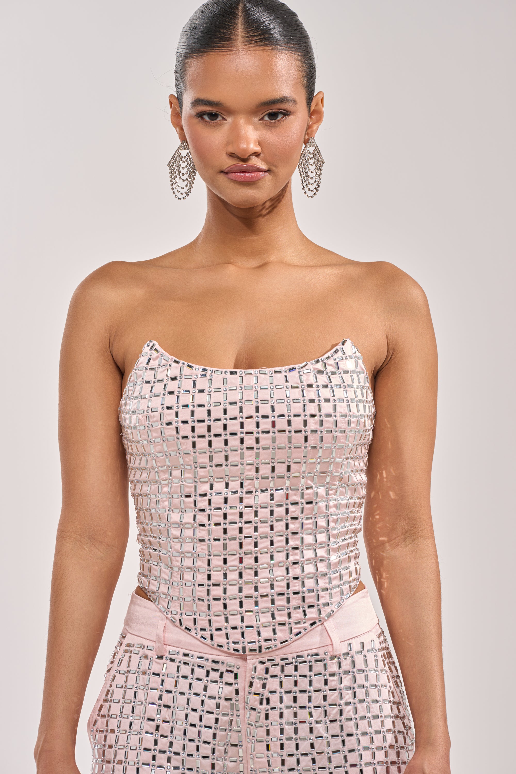 A woman wears the LILA EMBELLISHED SATIN CORSET IN LIGHT PINK, a strapless top with a silver geometric grid pattern, paired with large statement earrings and her hair pulled back against a plain background. 1