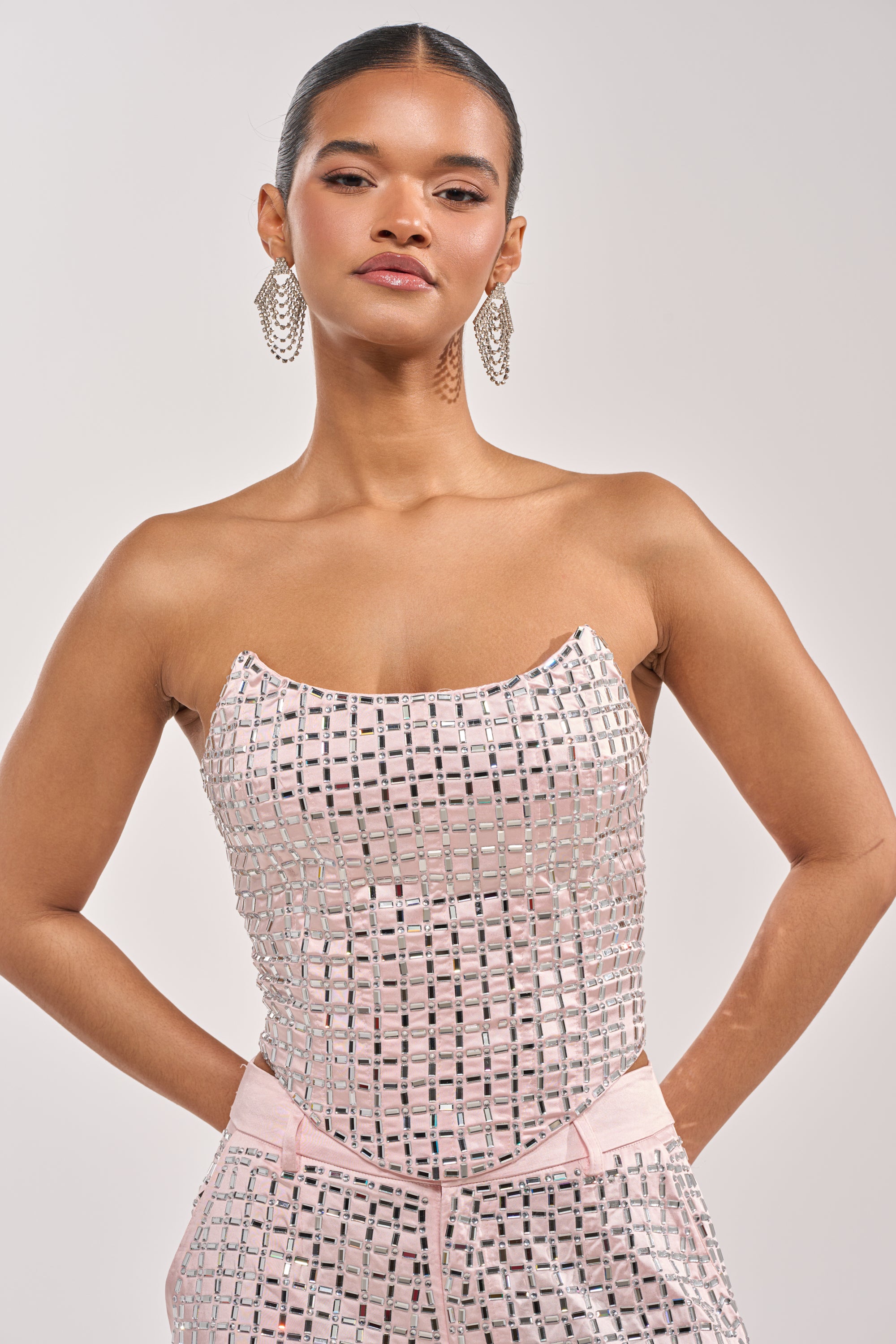 A woman poses confidently in the LILA EMBELLISHED SATIN CORSET IN LIGHT PINK with matching mirrored pants and bold geometric earrings. Her hair is pulled back neatly as she stands against a plain background. 2
