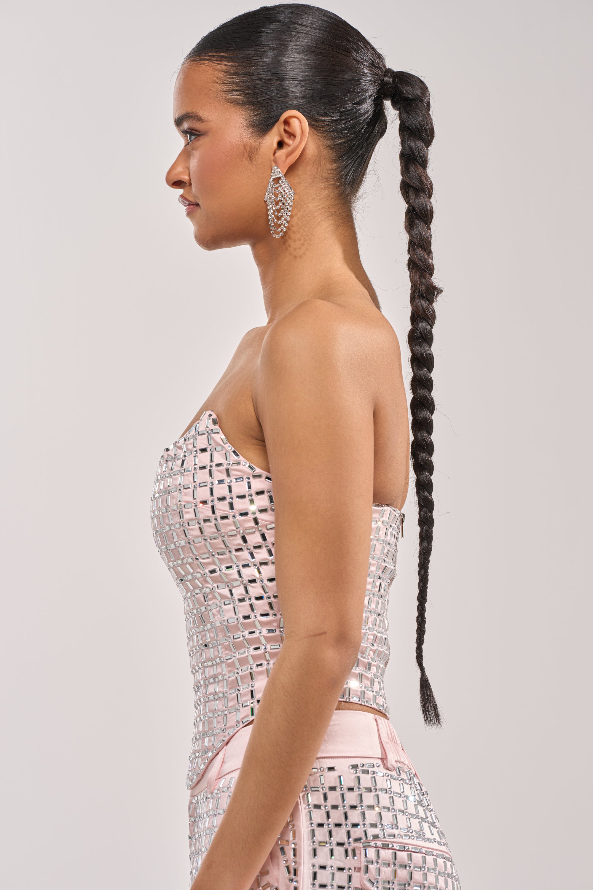 A woman in profile with a long braid wears the LILA EMBELLISHED SATIN CORSET IN LIGHT PINK with large dangling earrings, standing against a plain light background. 3