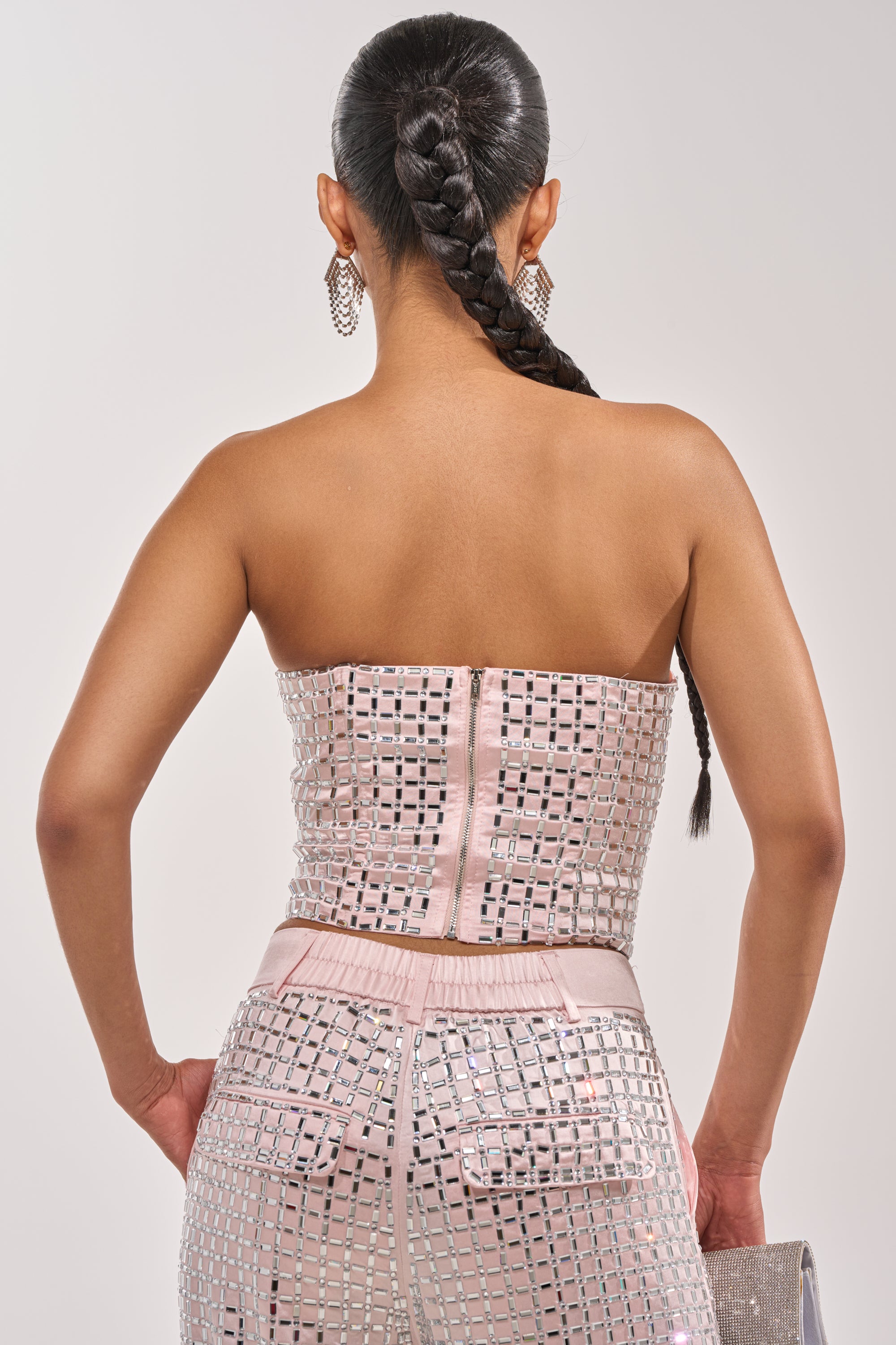 A woman with a long braided ponytail is shown from the back wearing the LILA EMBELLISHED SATIN CORSET IN LIGHT PINK, adorned with square mirrors and paired with a glittery silver clutch and large sparkling earrings. 4