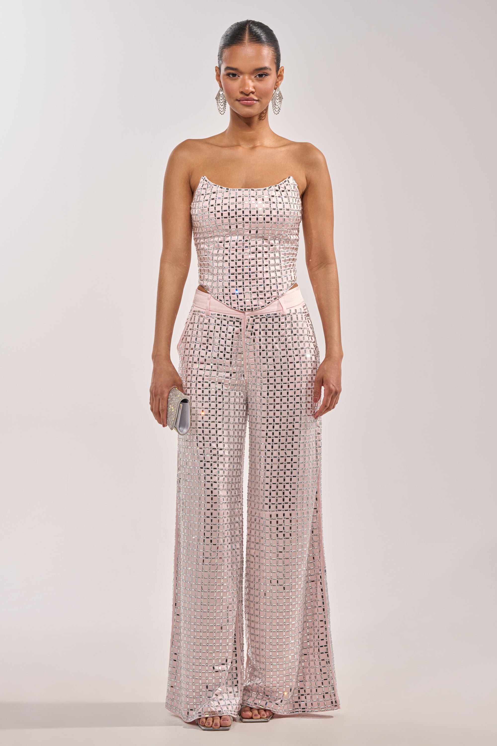 A woman faces forward in the LILA EMBELLISHED SATIN CORSET IN LIGHT PINK, styled with a sequin jumpsuit, wide legs, small clutch, drop earrings, and slicked-back hair. 5