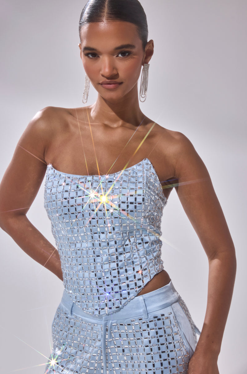 A woman poses confidently in the LILA EMBELLISHED SATIN CORSET and matching pants, her slicked-back hair and statement earrings shining as starbursts of light reflect off her square-patterned, sequin-embellished outfit.