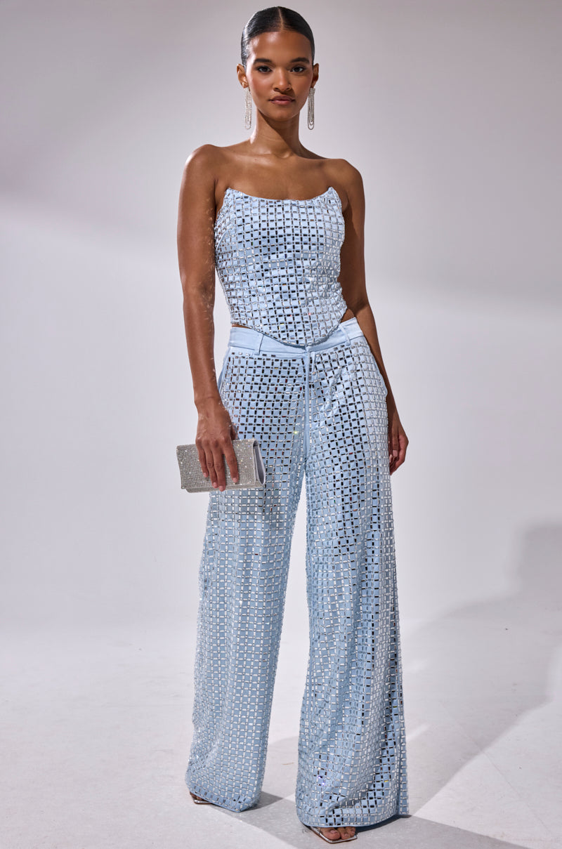A woman poses against a plain background wearing the LILA EMBELLISHED SATIN CORSET, paired with wide-leg trousers. She accessorizes with a small silver clutch, drop earrings, and has her hair styled sleekly back.