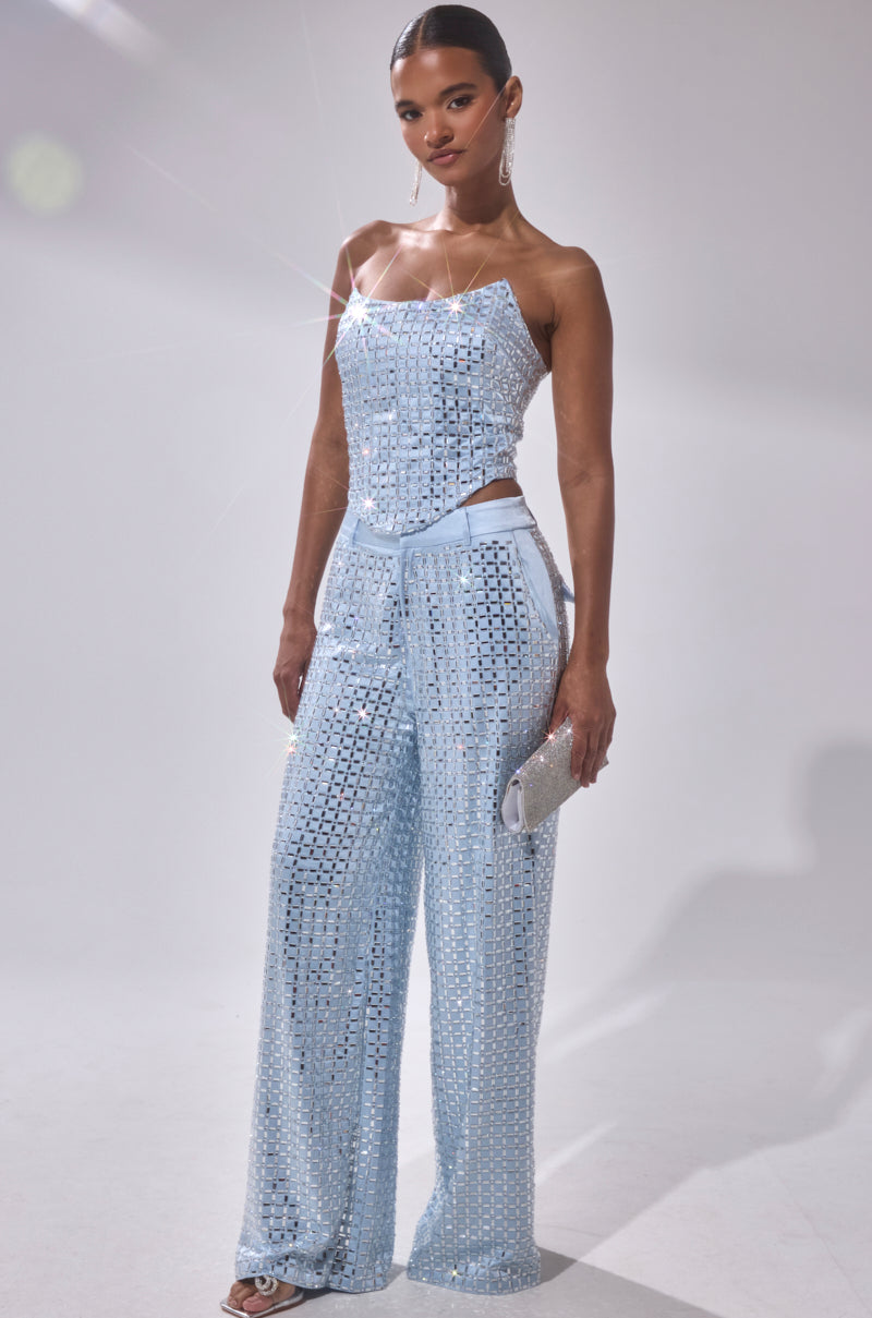 A woman poses against a plain white background in the LILA EMBELLISHED SATIN CORSET and wide-leg pants, holding a silver clutch and wearing dangling earrings. The shimmering light blue outfit features sequins and a strapless top.