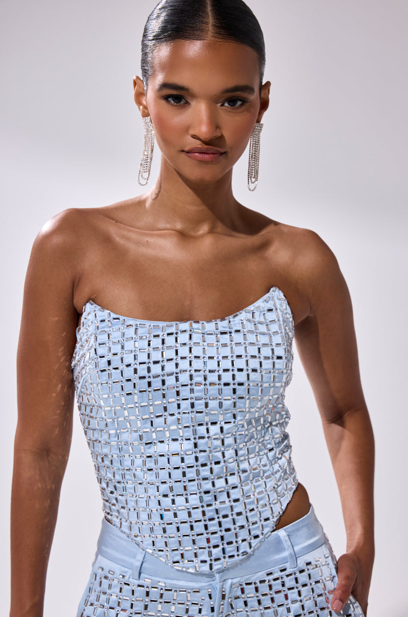 A woman with slicked-back hair wears long, dangling earrings and the LILA EMBELLISHED SATIN CORSET in light blue with a grid pattern, paired with matching pants, standing against a plain background.