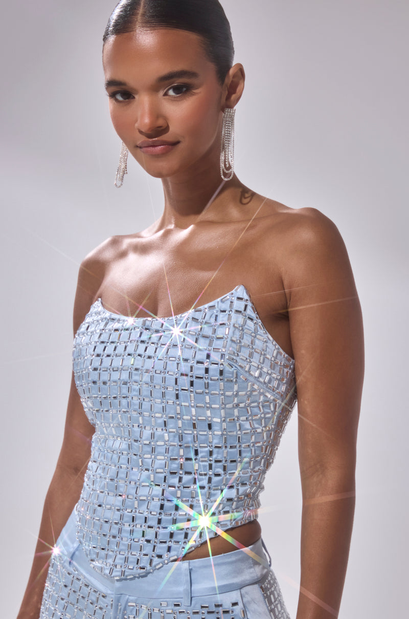 A woman with slicked-back hair models the LILA EMBELLISHED SATIN CORSET, a sparkly light blue top with square patterns and star-shaped reflections, paired with dangling earrings against a light background.