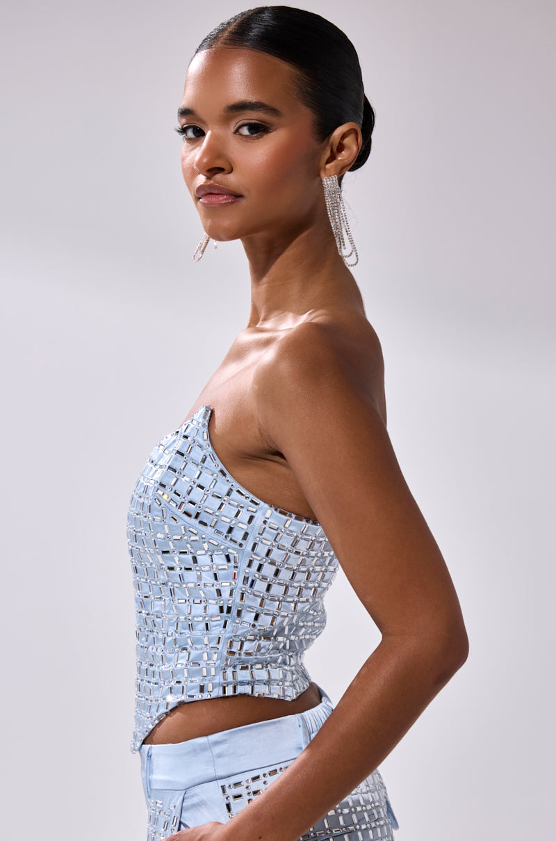 A woman with slicked-back hair in a side bun models the LILA EMBELLISHED SATIN CORSET, facing sideways toward the camera. She pairs it with matching pants and long, dangling earrings against a plain light gray background.