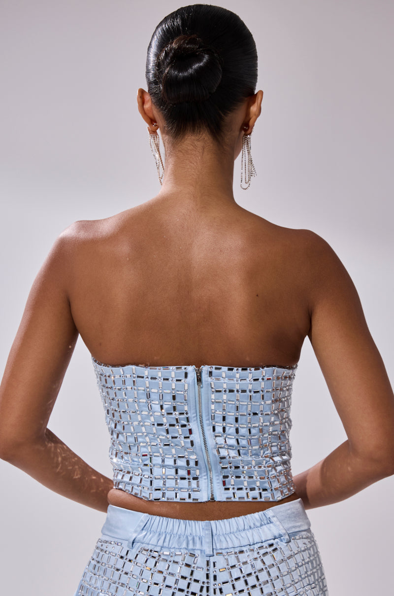 A woman with dark hair in a sleek bun and dangling earrings is seen from behind, wearing the LILA EMBELLISHED SATIN CORSET—a light blue, strapless top with a grid pattern, visible zipper, and matching high-waisted bottoms.