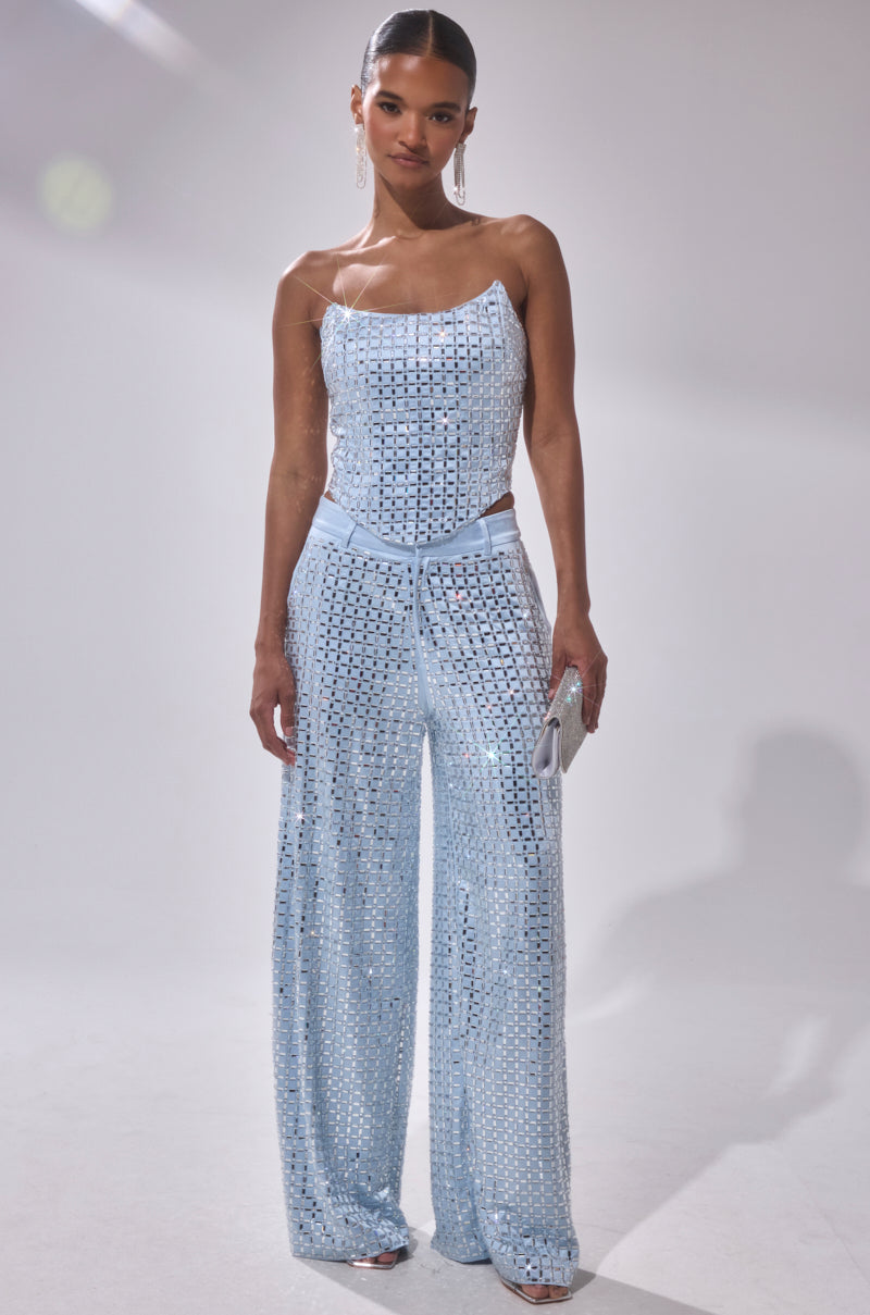 A woman stands against a white background wearing the LILA EMBELLISHED SATIN CORSET, a light blue, sequined, sleeveless top paired with matching wide-leg pants. She holds a silver clutch and accessorizes with long earrings and pulled-back hair.