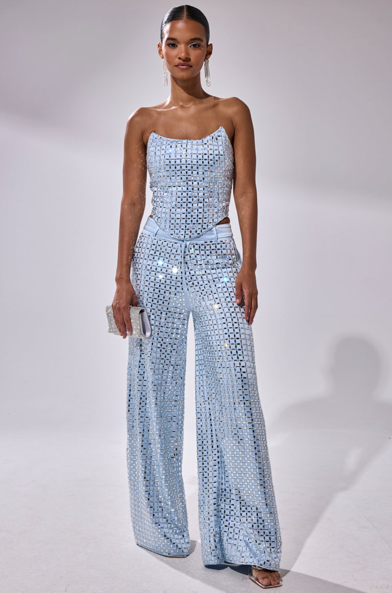 A woman stands against a plain background wearing the LILA EMBELLISHED SATIN CORSET, styled with wide-leg pants, a small clutch, dangling earrings, and open-toe heels.