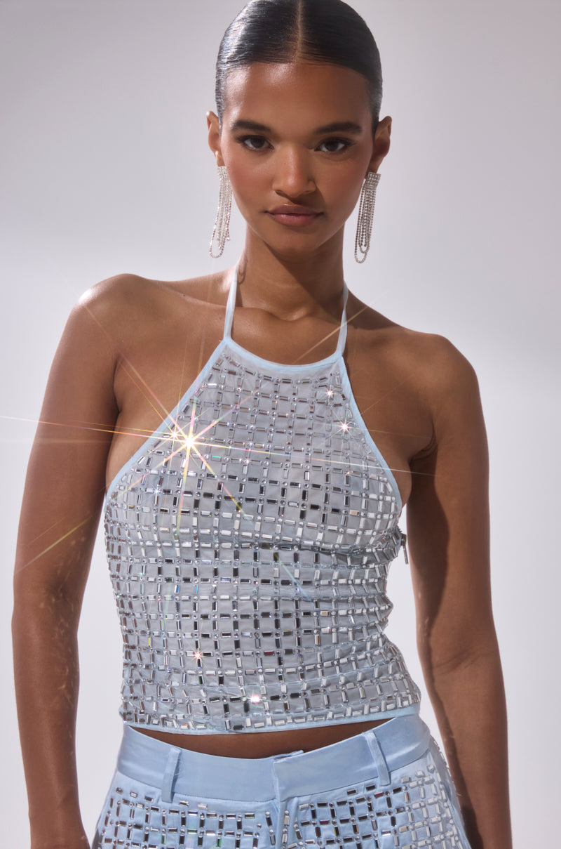 A woman stands confidently in the LILA EMBELLISHED MOCK NECK MESH TOP, a powder blue crop top with reflective square mirror embellishments, her hair in a sleek bun and long earrings highlighting her subtle smile.