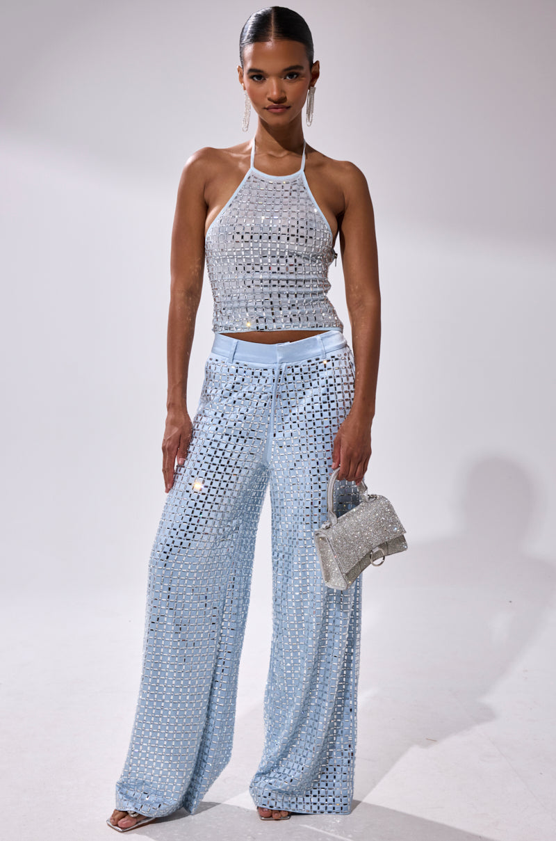 A woman stands confidently in the LILA EMBELLISHED MOCK NECK MESH TOP paired with high-waisted, wide-leg pants, holding a glittery silver handbag. Her dark hair is styled in a sleek, middle-parted bun.