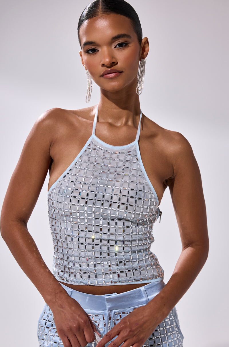 A woman poses confidently in the LILA EMBELLISHED MOCK NECK MESH TOP, featuring silver square sequins, styled with light blue pants. She completes the look with slicked-back hair and large earrings against a plain background.