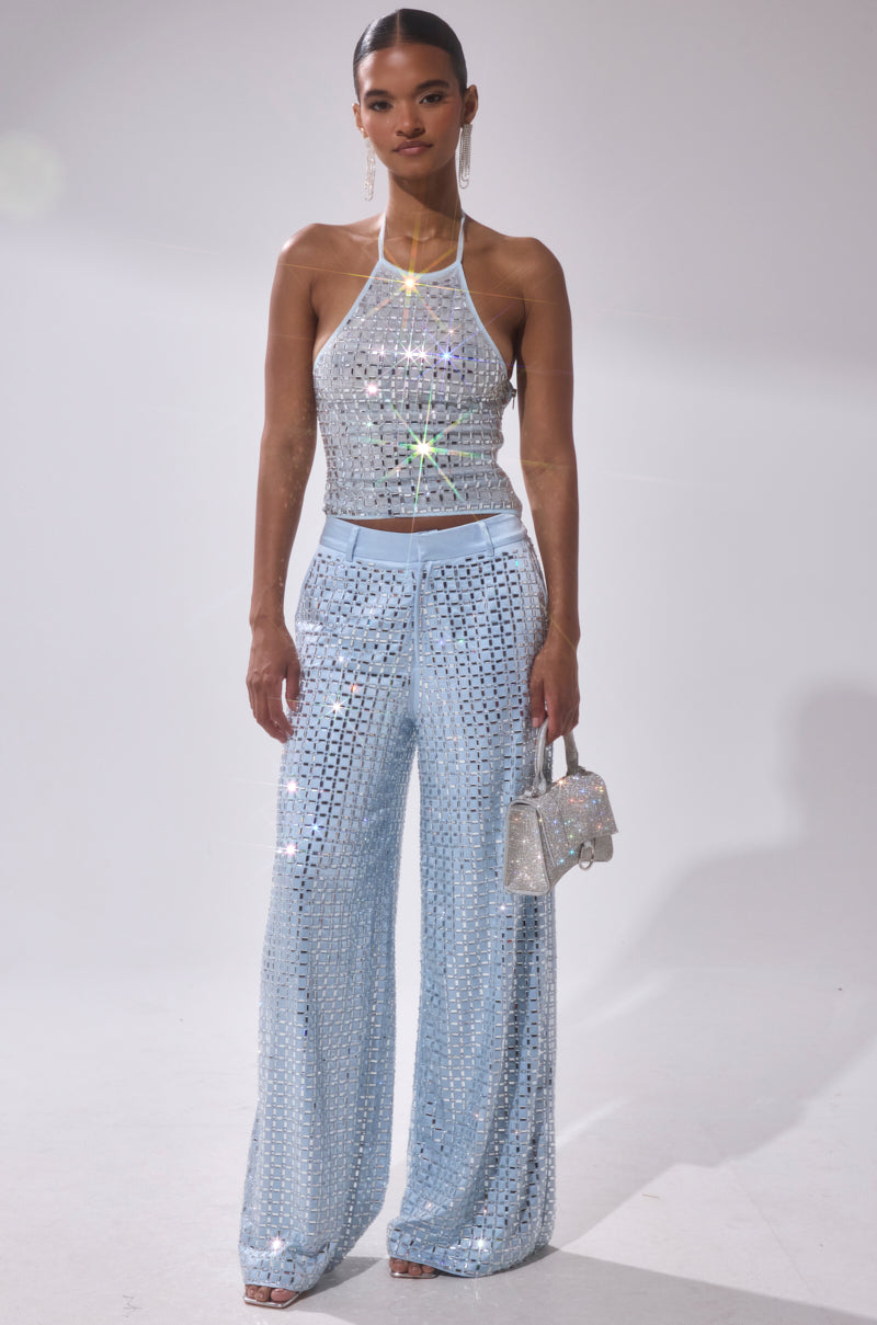 A woman poses against a plain background in the LILA EMBELLISHED MOCK NECK MESH TOP, a shimmering powder blue crop top with sequins, paired with matching wide-leg pants and a glittery handbag. Her hair is pulled back for a polished effect.