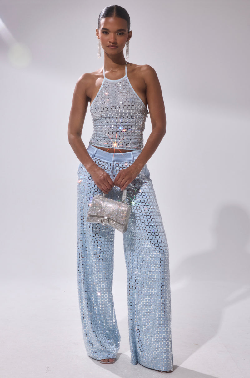 A woman stands against a light background wearing the LILA EMBELLISHED MOCK NECK MESH TOP with matching wide-leg pants, holding a glittery silver clutch. She has sleek hair, subtle makeup, and large dangling earrings.