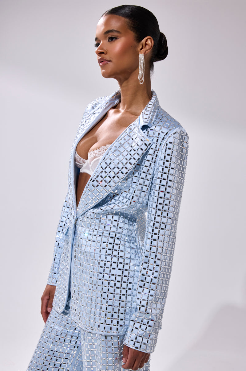 A woman poses against a light background wearing the LILA EMBELLISHED SATIN BLAZER in shiny light blue with grid patterns and mirrored embellishments, layered over a white lace bra. She accessorizes with long earrings and a sleek bun.