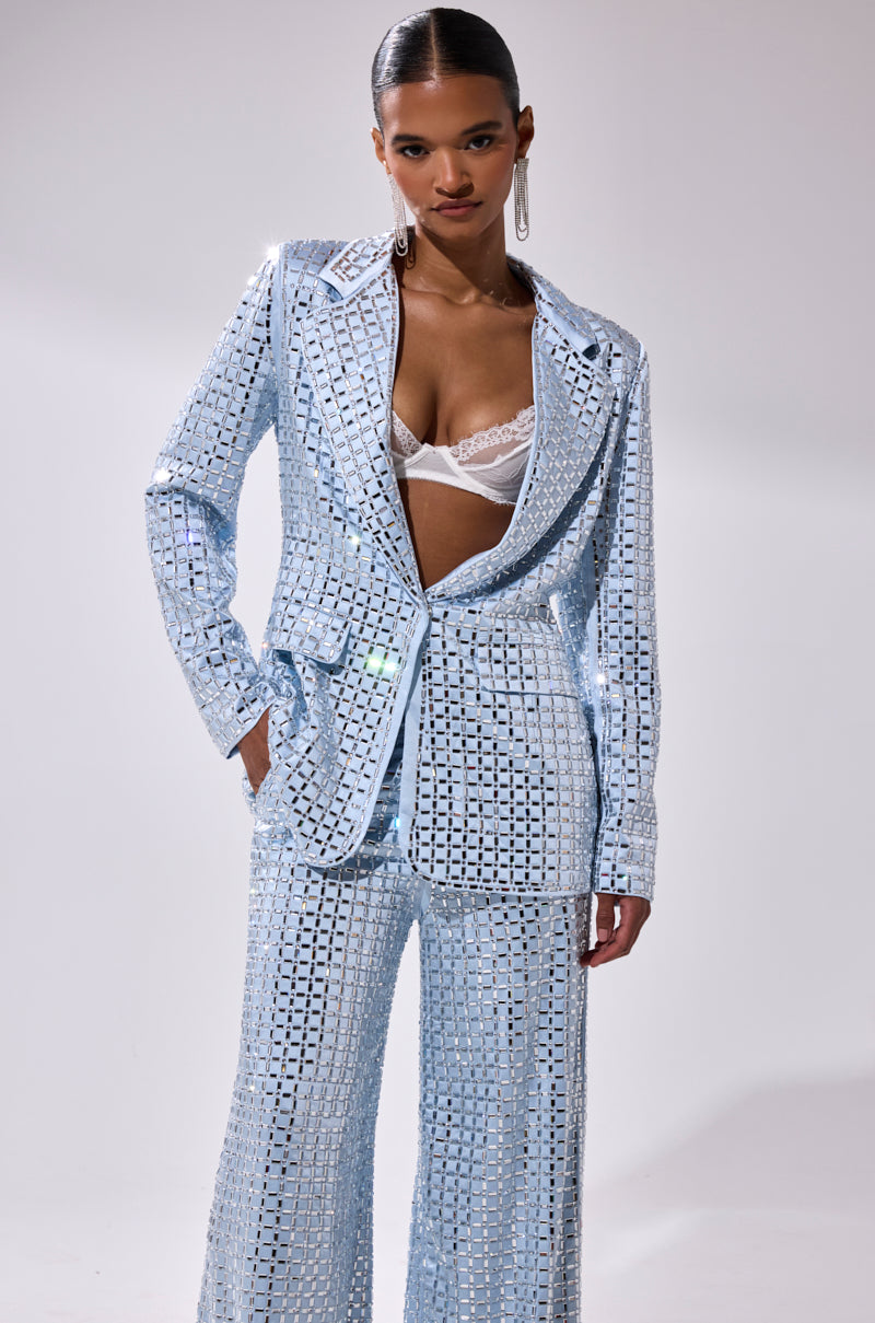 A woman poses in a shiny, light blue LILA EMBELLISHED SATIN BLAZER with a deep V-neck that reveals a white lace bra. She has sleek, pulled-back hair and wears large, dangling earrings.