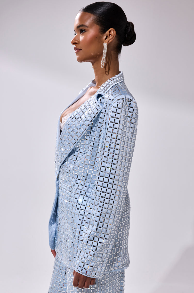 A woman stands in profile against a plain background, wearing the light blue AKIRA Label LILA EMBELLISHED SATIN BLAZER with grid patterns and square cutouts. Statement earrings and a sleek bun highlight her geometric mirrored details.